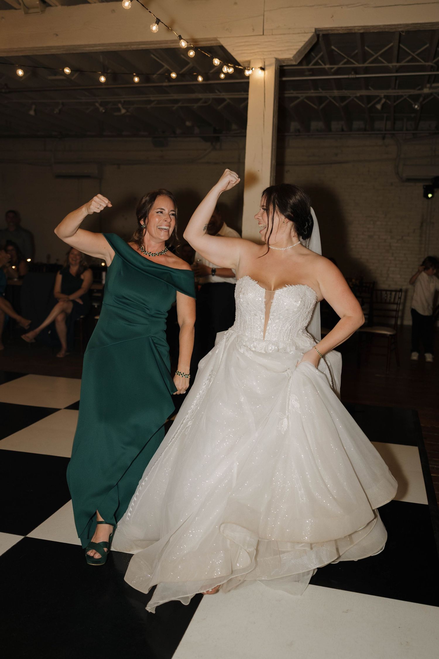 Bride in white ballgown dances joyfully with wedding party members in emerald green dresses during reception.