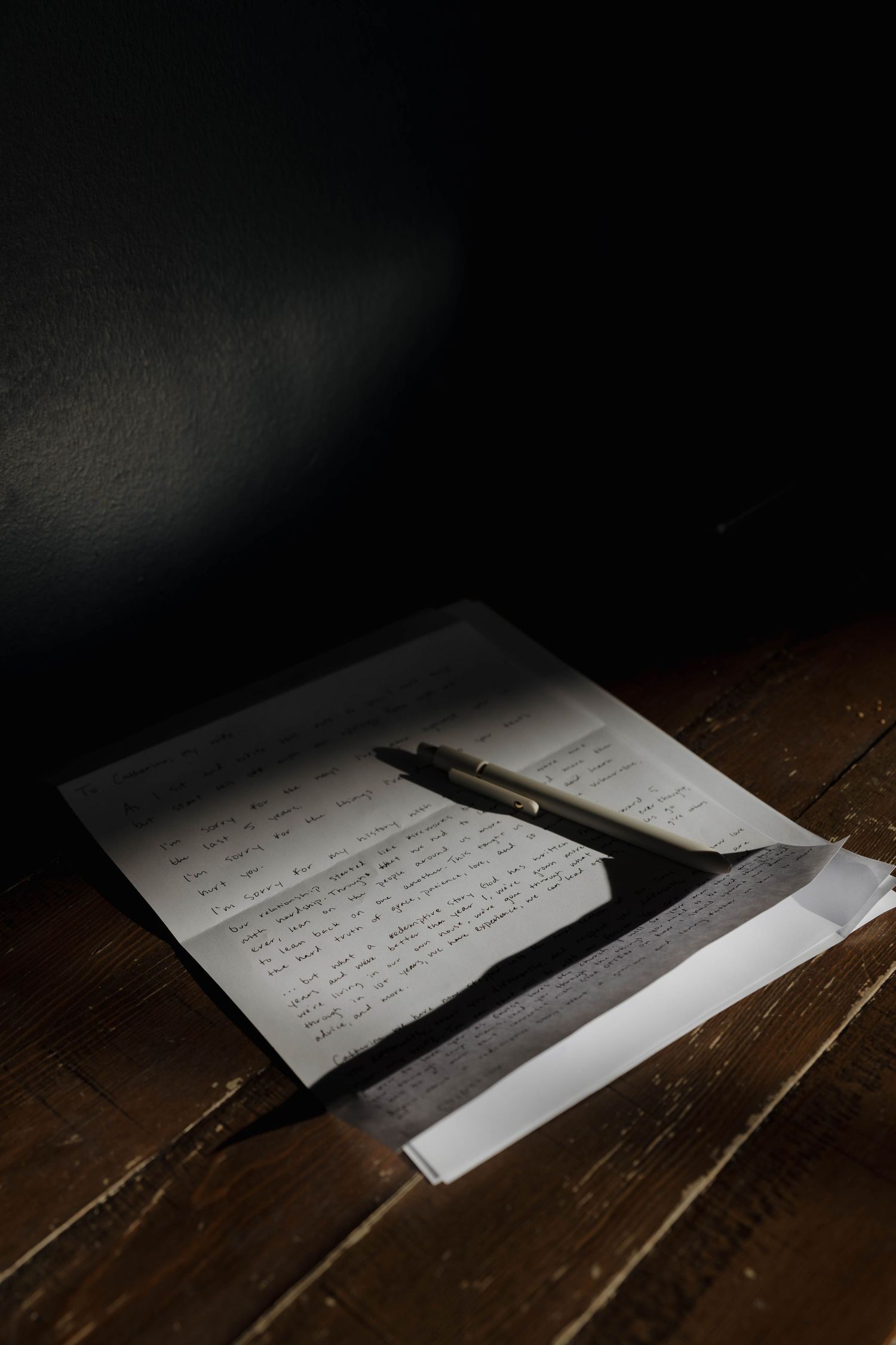 Handwritten letter on dark wooden table with dramatic lighting and a pen resting on the paper.