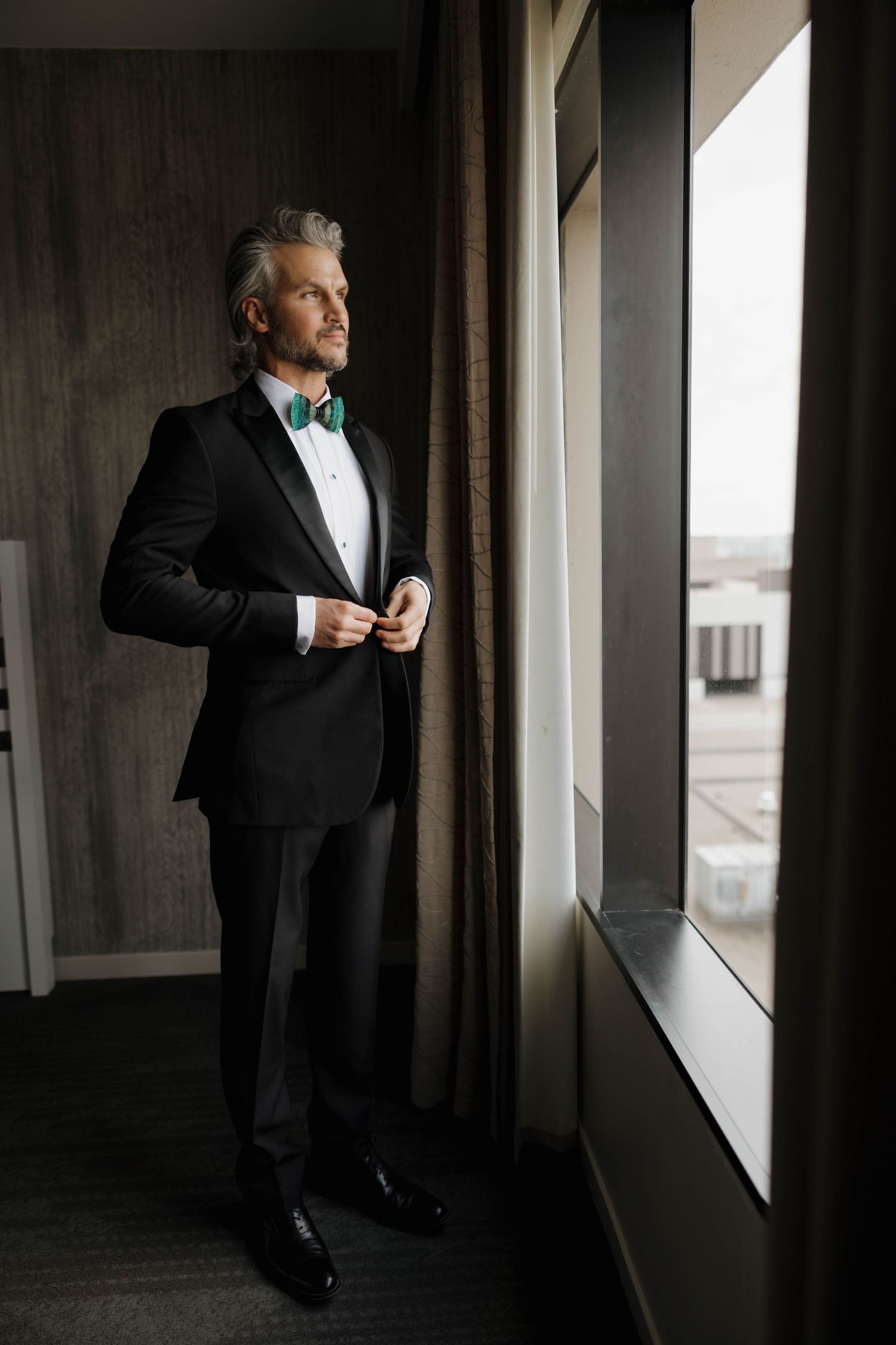 A distinguished person in a black tuxedo and turquoise bow tie gazes contemplatively out a window.
