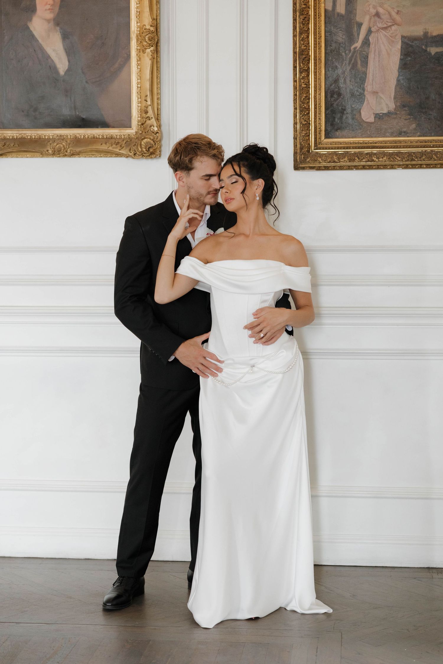 A couple shares an intimate moment in an elegant white-walled room with ornate gold-framed paintings.