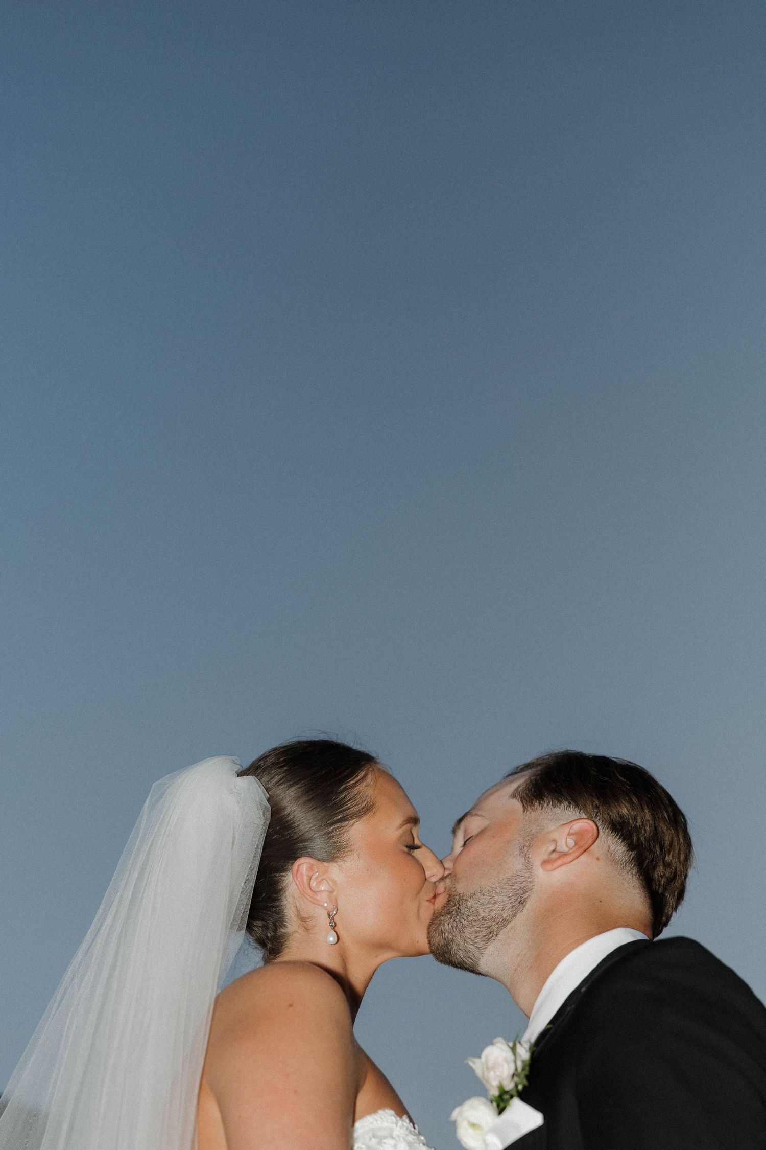 Newlyweds share an intimate kiss against a blue sky backdrop while wearing traditional wedding attire.
