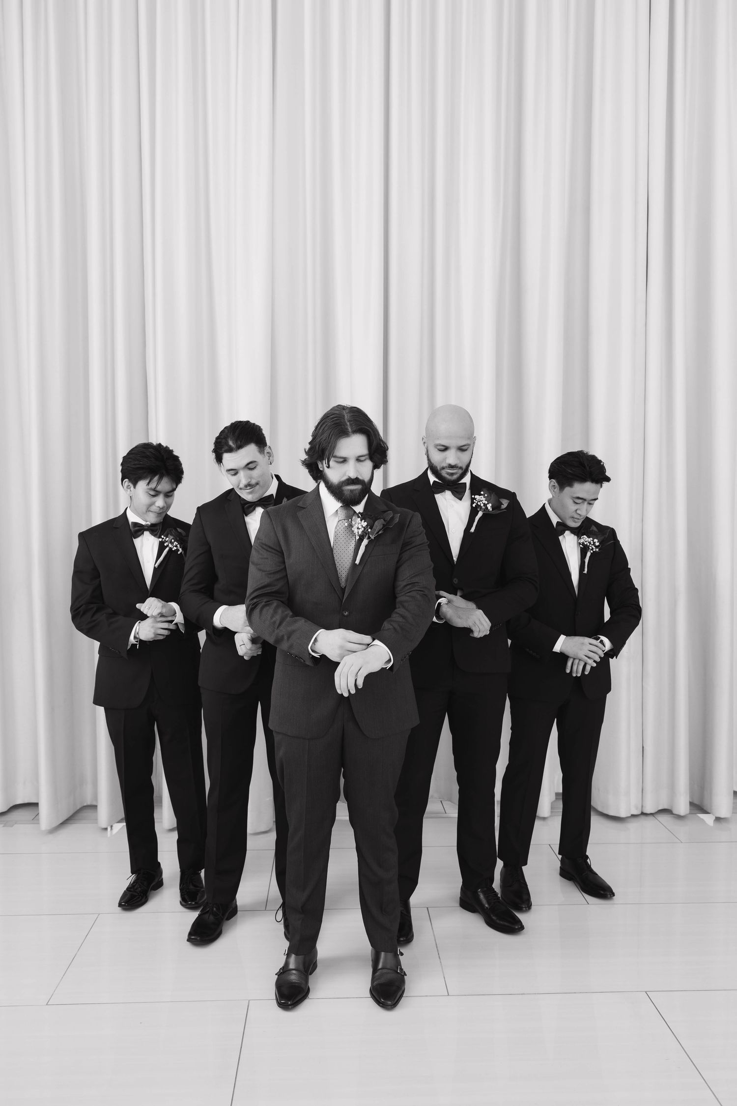 A black and white photo shows several men in suits sitting on the floor against a white curtain background.