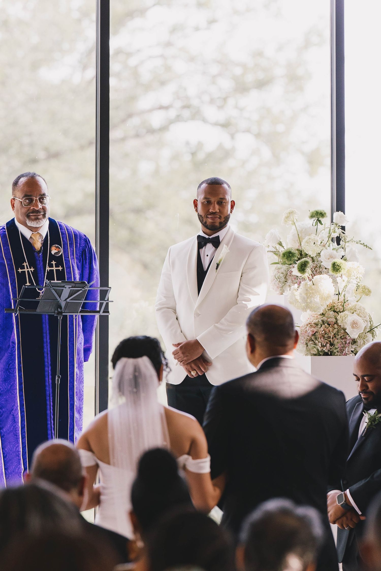A wedding ceremony takes place with clergy in purple robes and groomsmen in white suits standing at the altar.