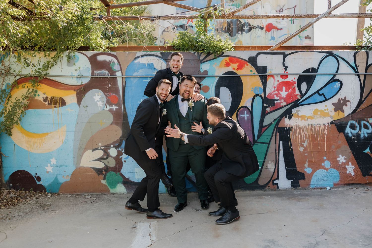 A group stands together posing playfully in front of a colorful graffiti wall on an urban street.