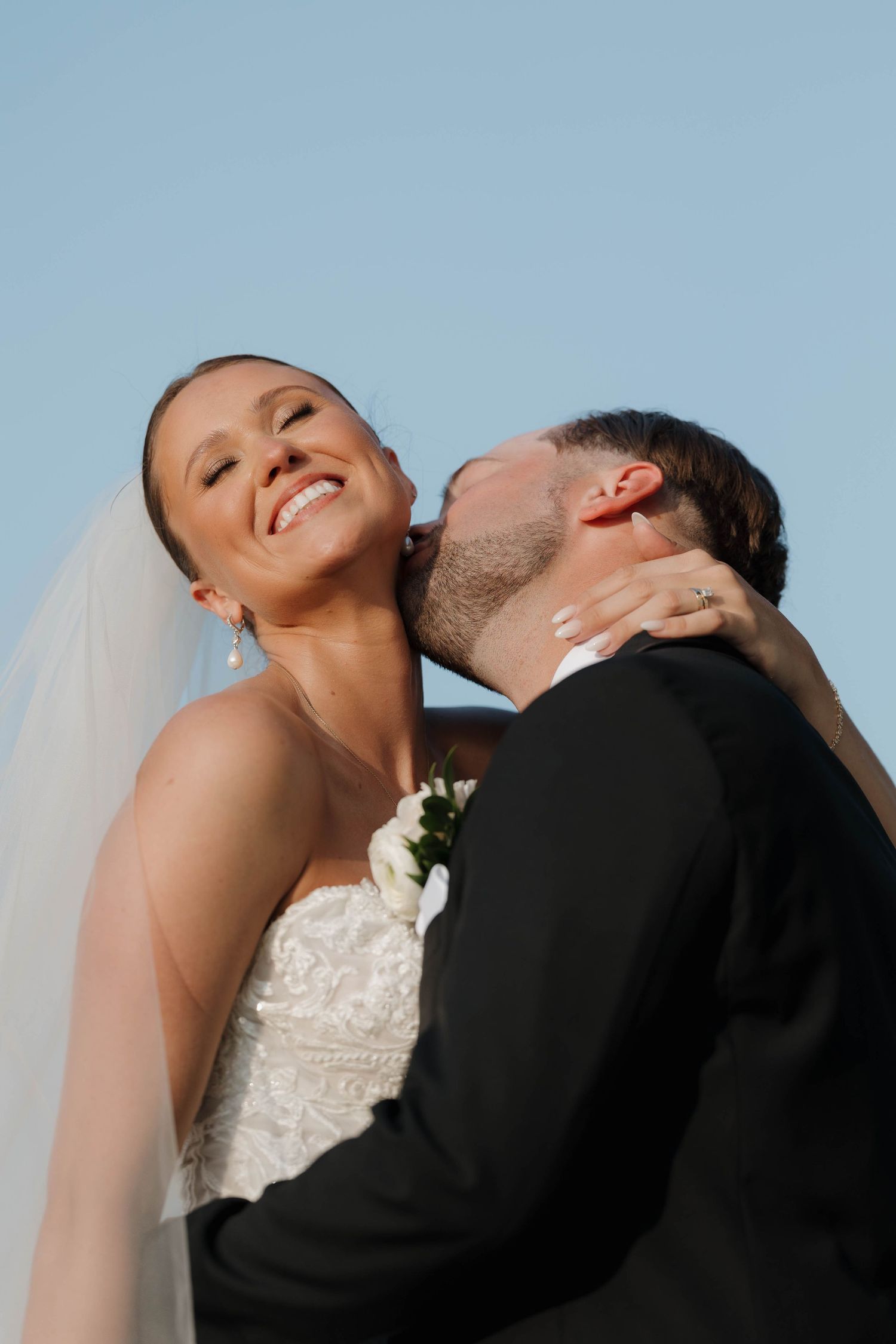 A bride in a white wedding dress leans back dramatically with her smiling groom in a tender embrace against blue sky.