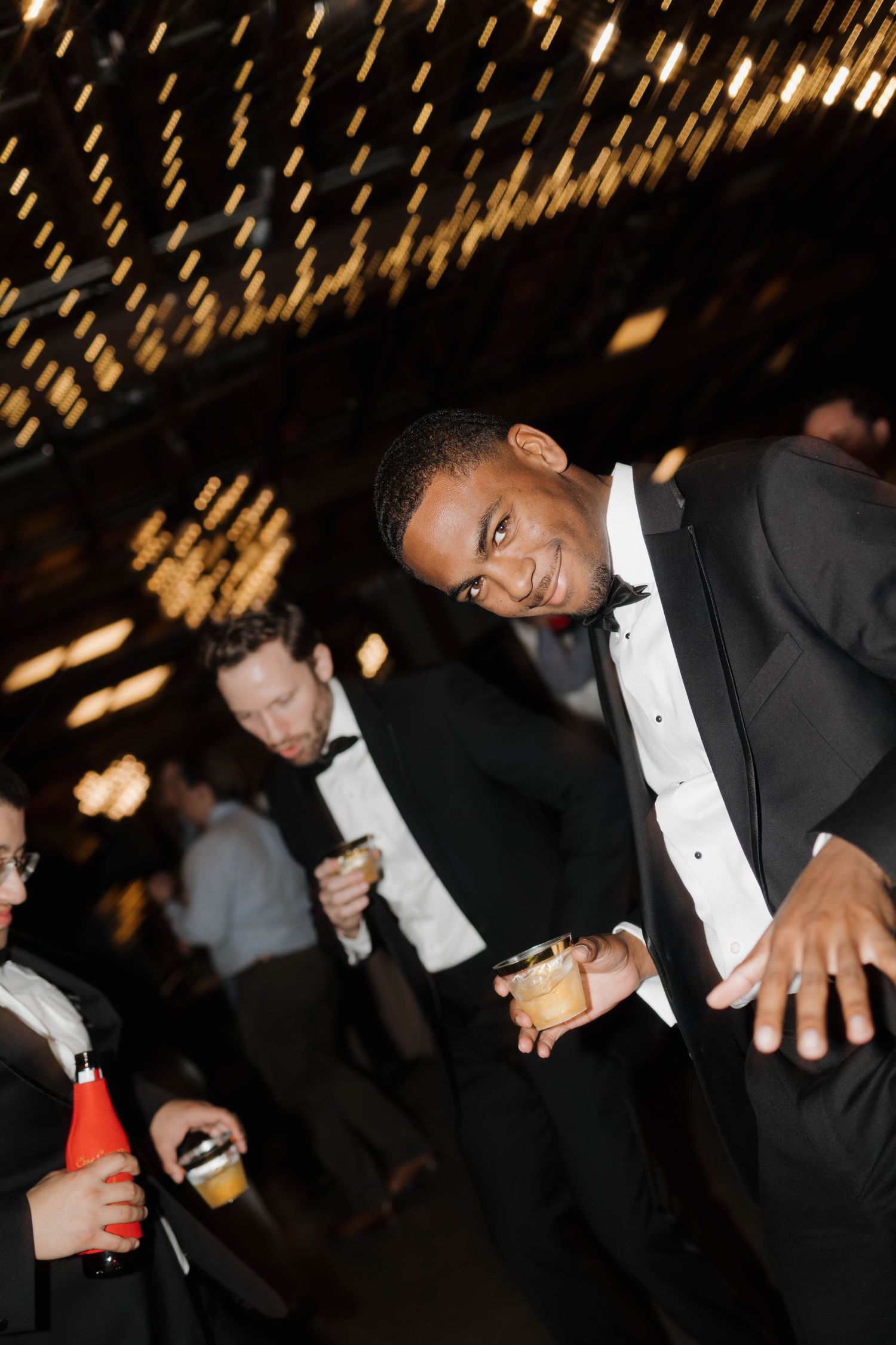 Wedding guests in formal black tie attire dancing and celebrating at an elegant reception with twinkling lights overhead.