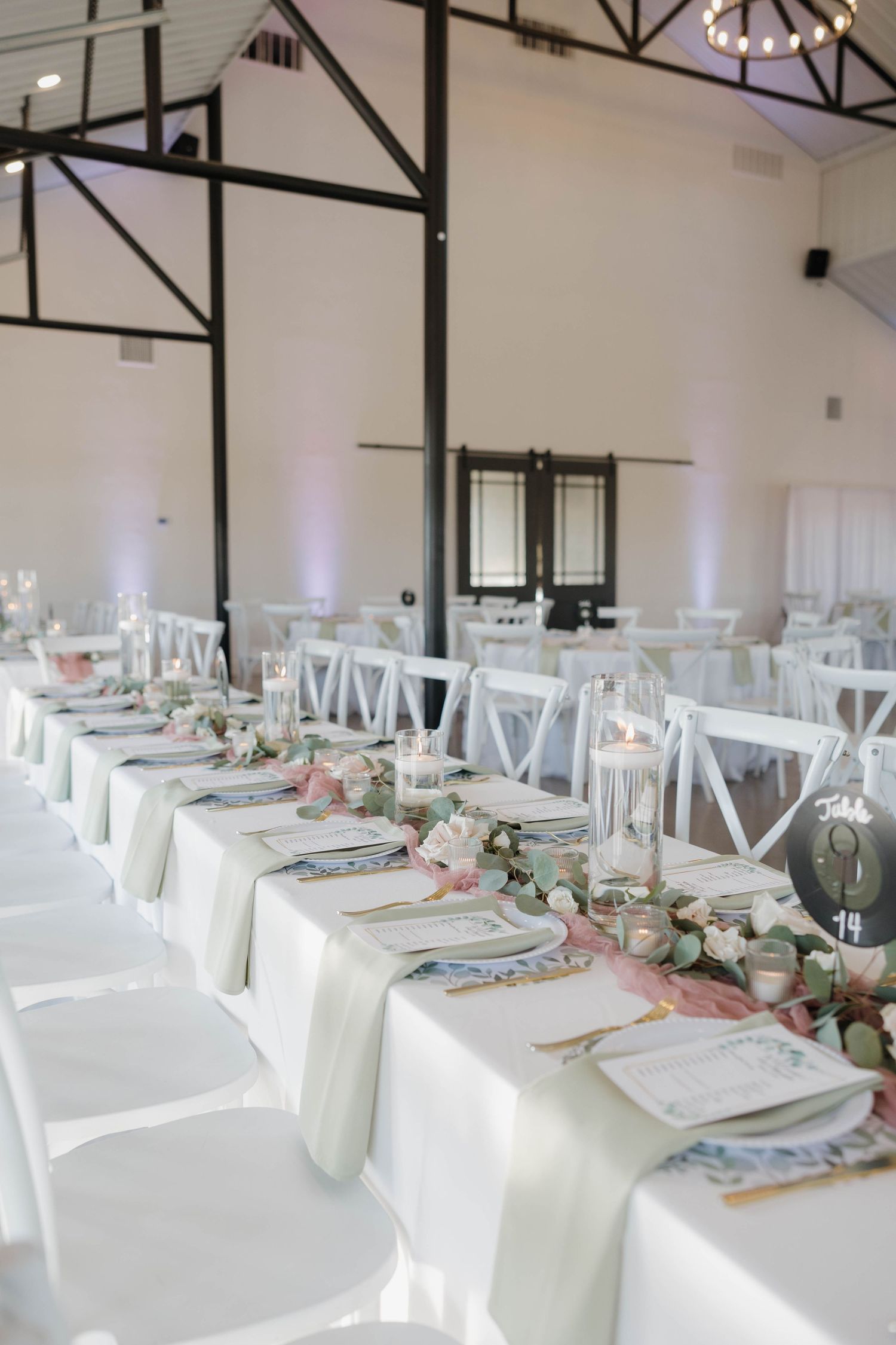 Elegant white reception tables with cross-back chairs set up in a bright indoor venue with rustic exposed beams.