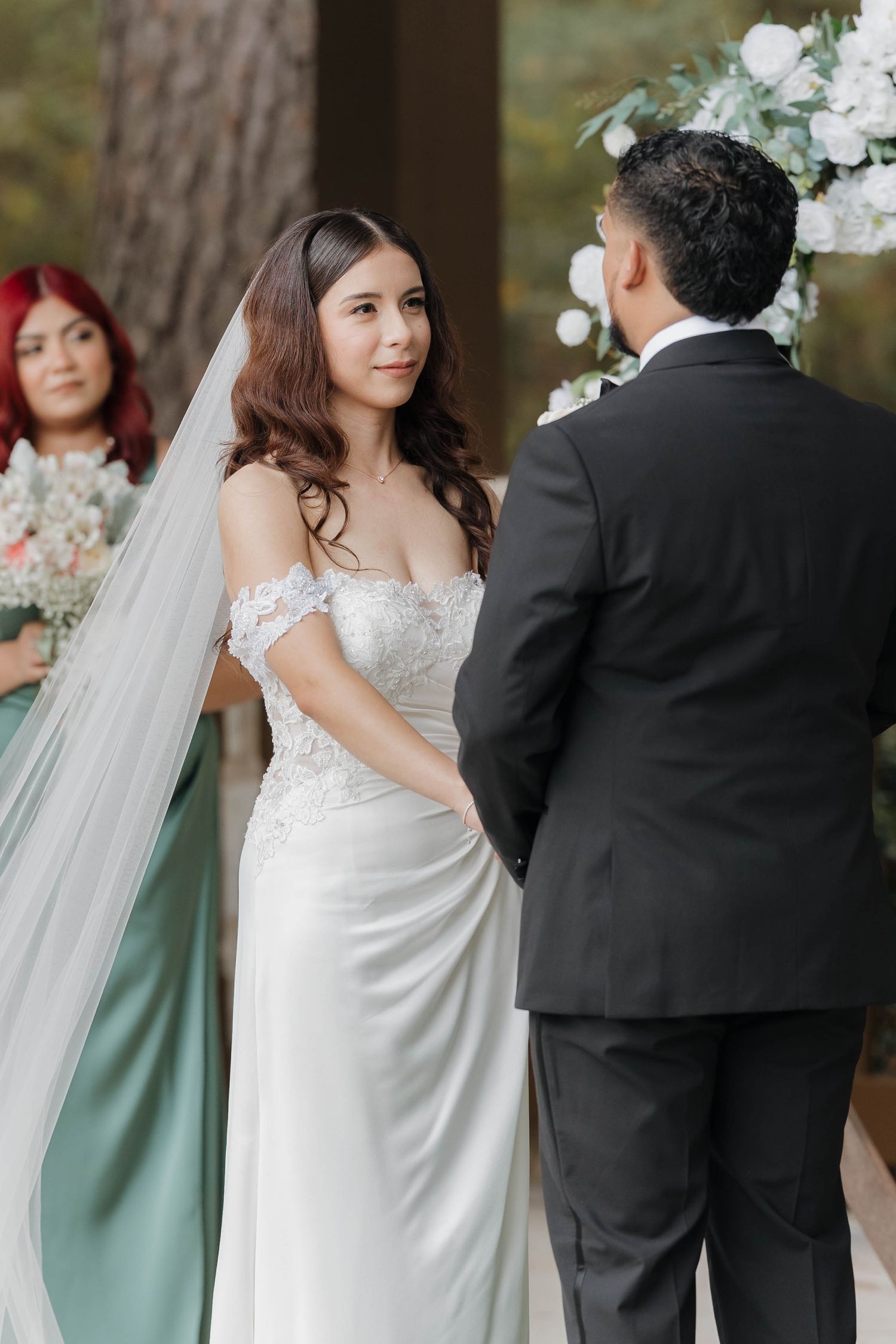 A bride in an elegant off-shoulder white gown stands with her groom during an outdoor wedding ceremony.