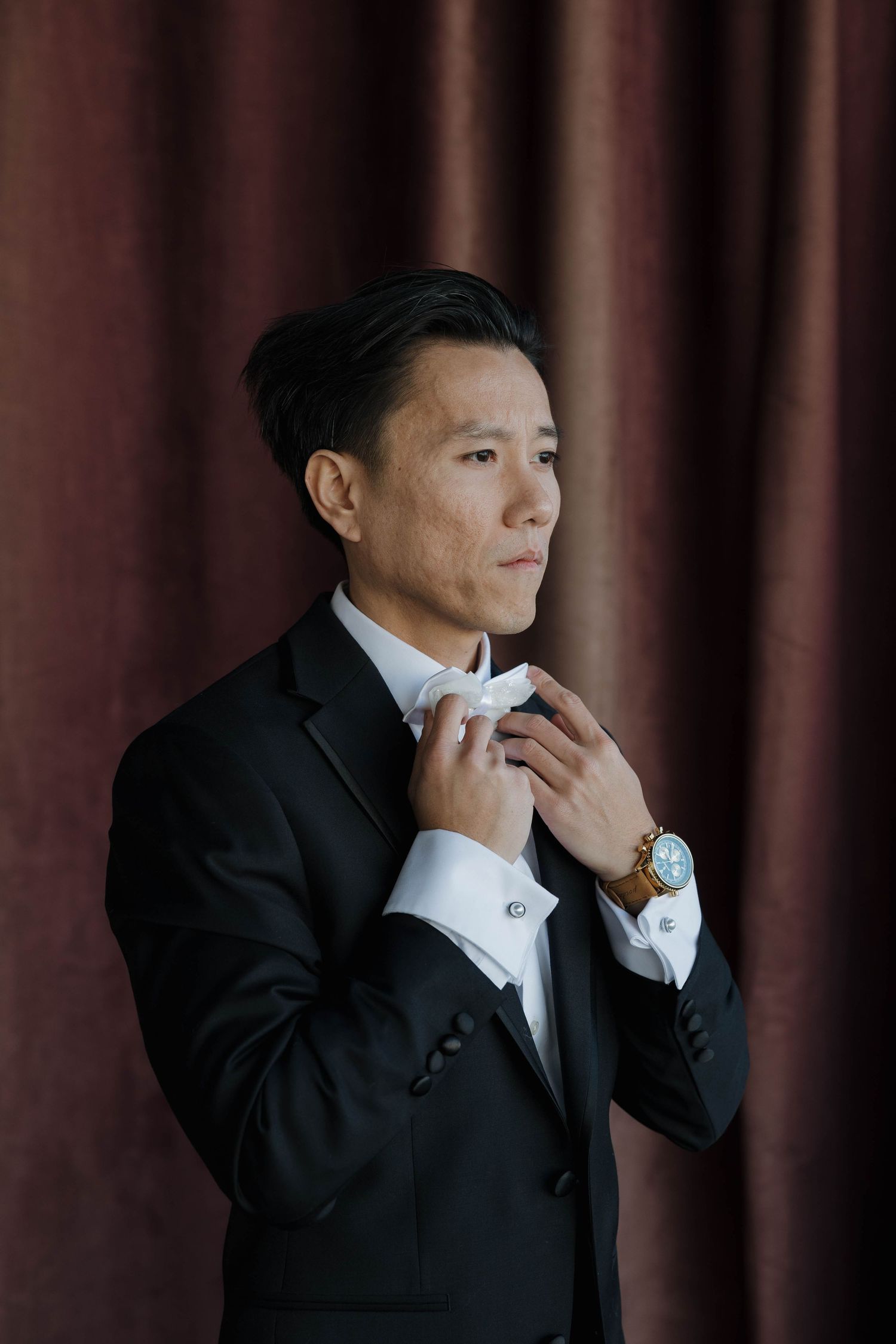 Close-up portrait of groomsmen in formal attire adjusting their bowties against a dark burgundy backdrop.