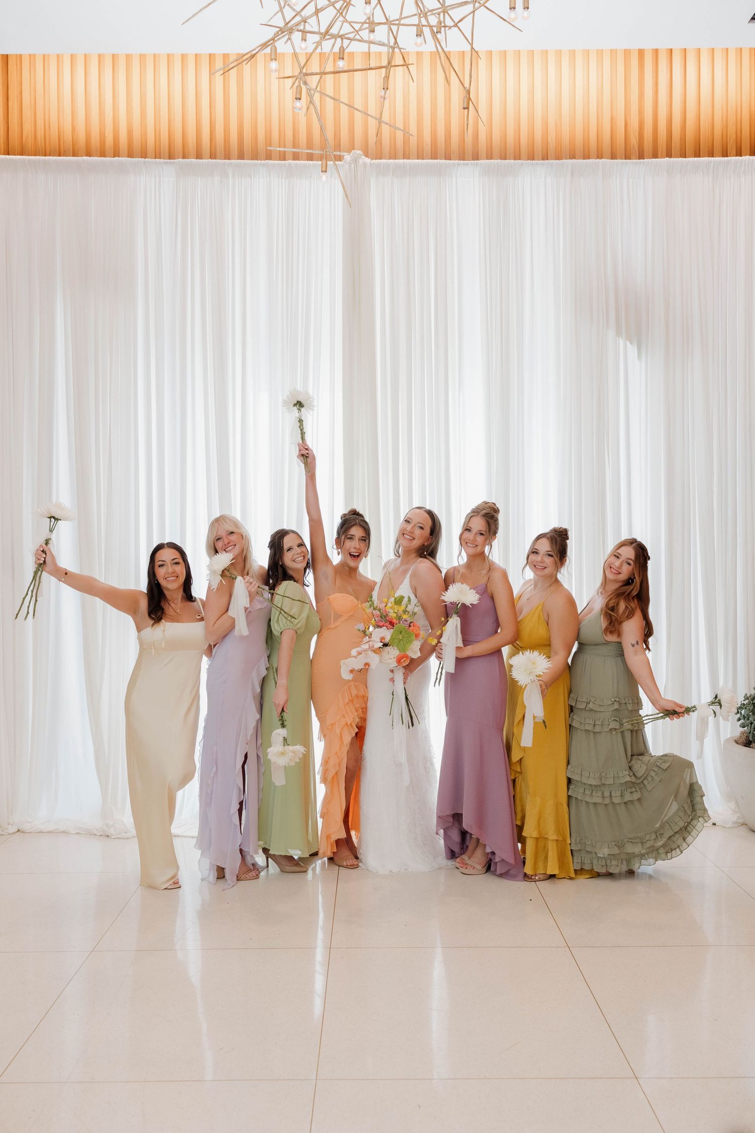 A group in pastel-colored dresses poses together holding bouquets in front of white curtains under an orange chandelier.
