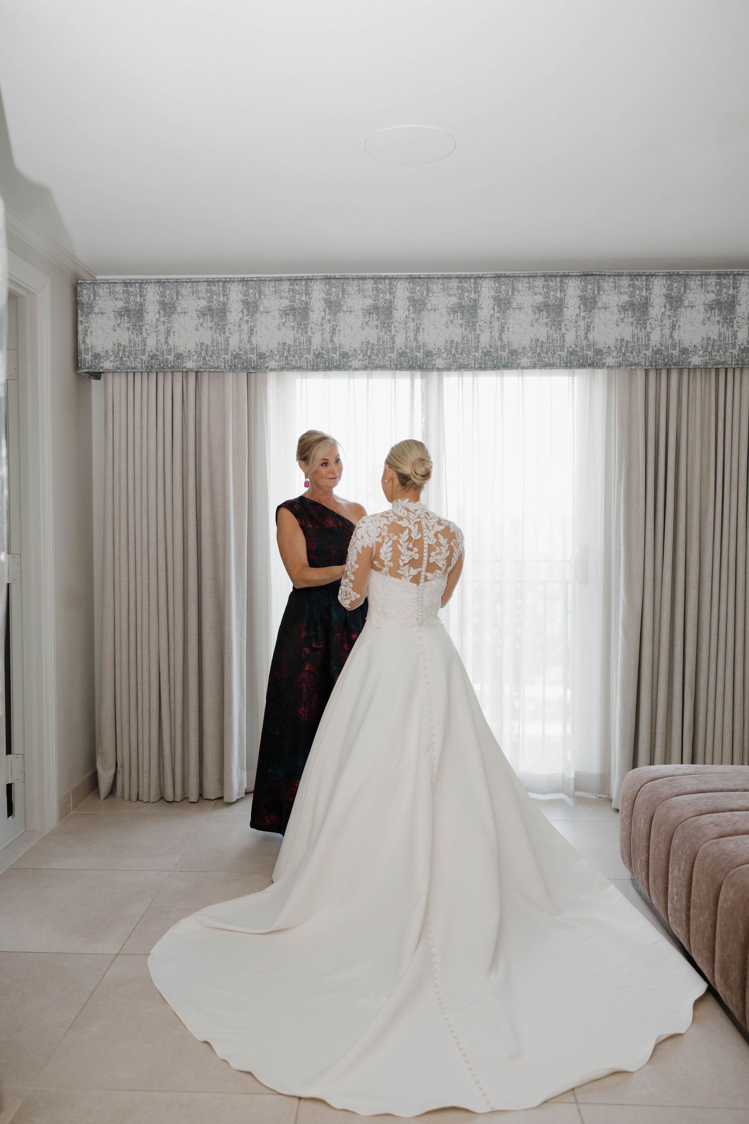 A wedding dress fitting session showing the elegant train of a white gown being adjusted in a bright room with beige curtains.