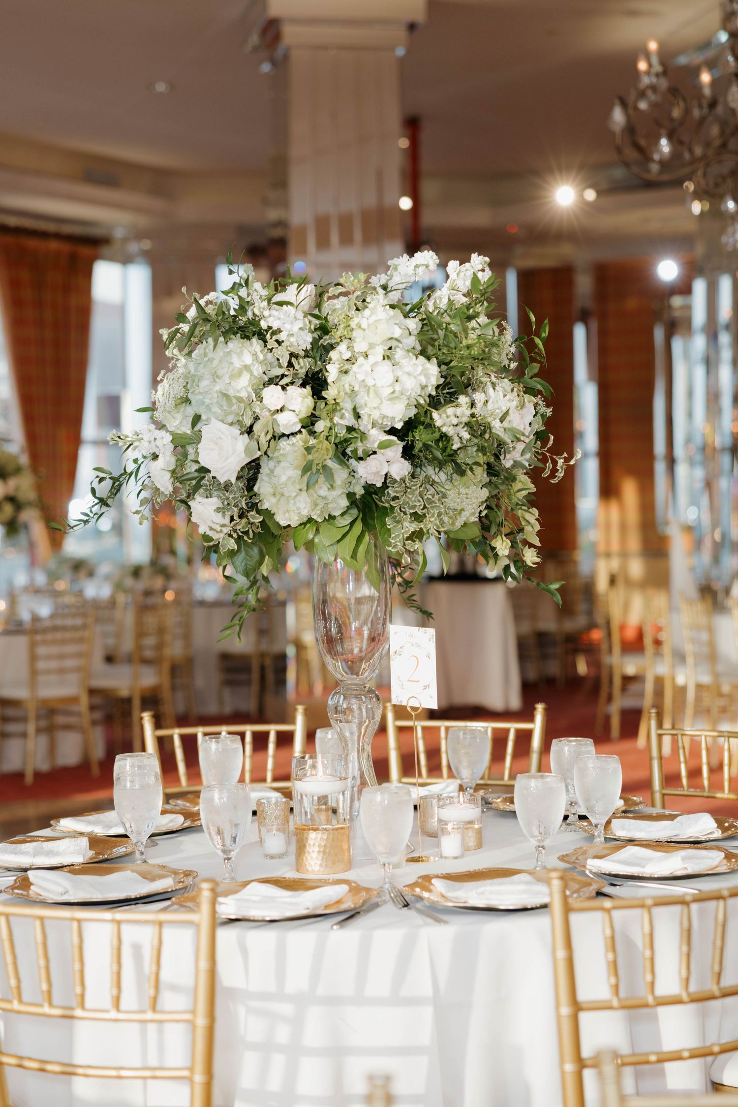 Luxurious wedding reception venue with white floral centerpieces and gold chiavari chairs at formal dining tables.