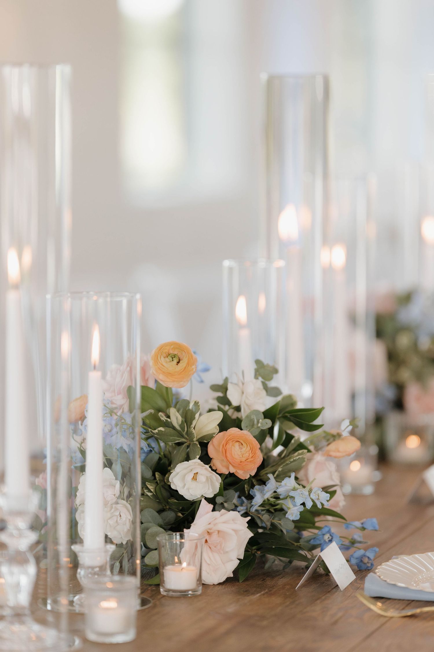 Elegant wedding table decor with tall glass candleholders, peach and white flowers, and eucalyptus greenery on wooden surface.