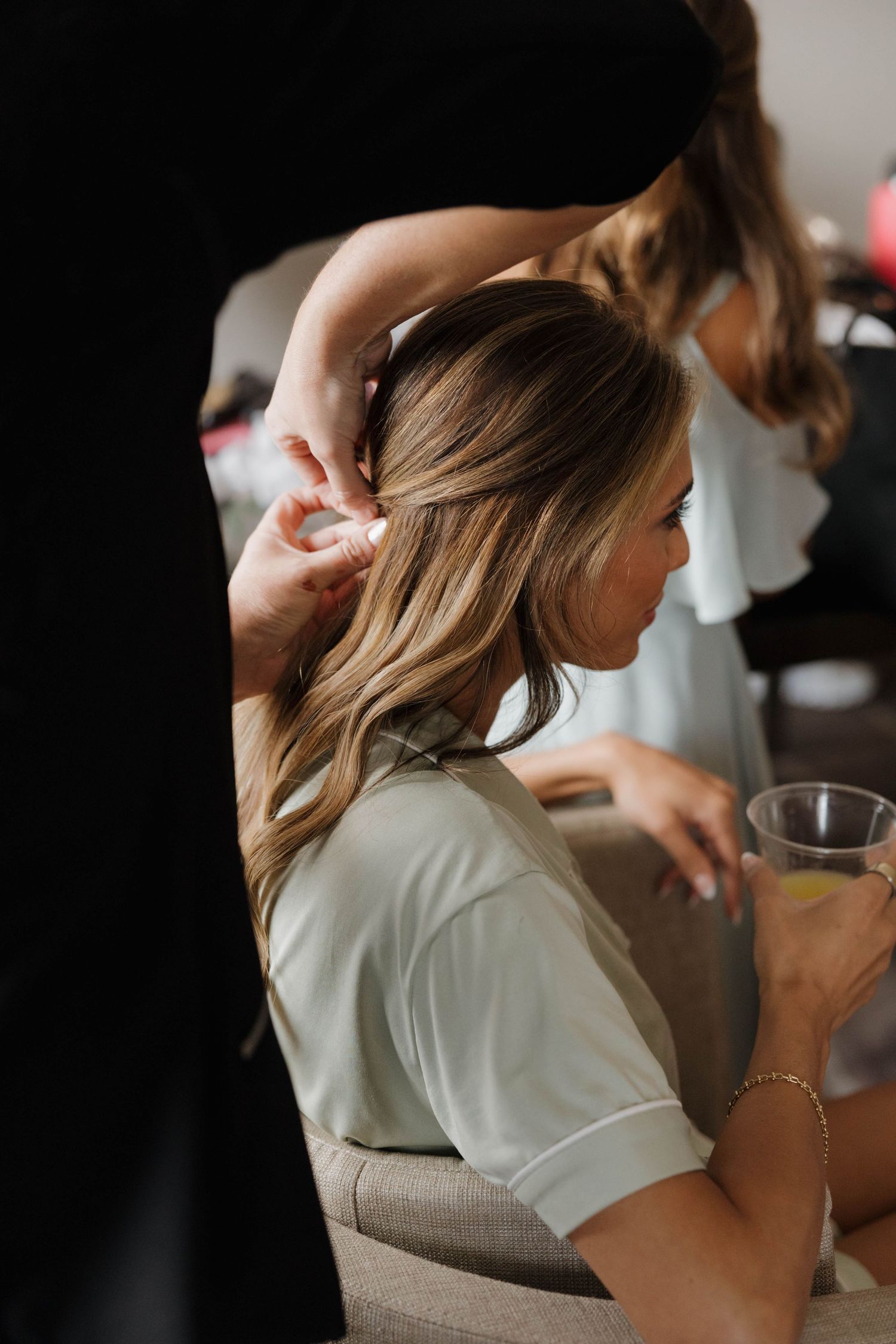 A woman in a white blouse gets her hair styled while holding a drink in a dimly lit setting.