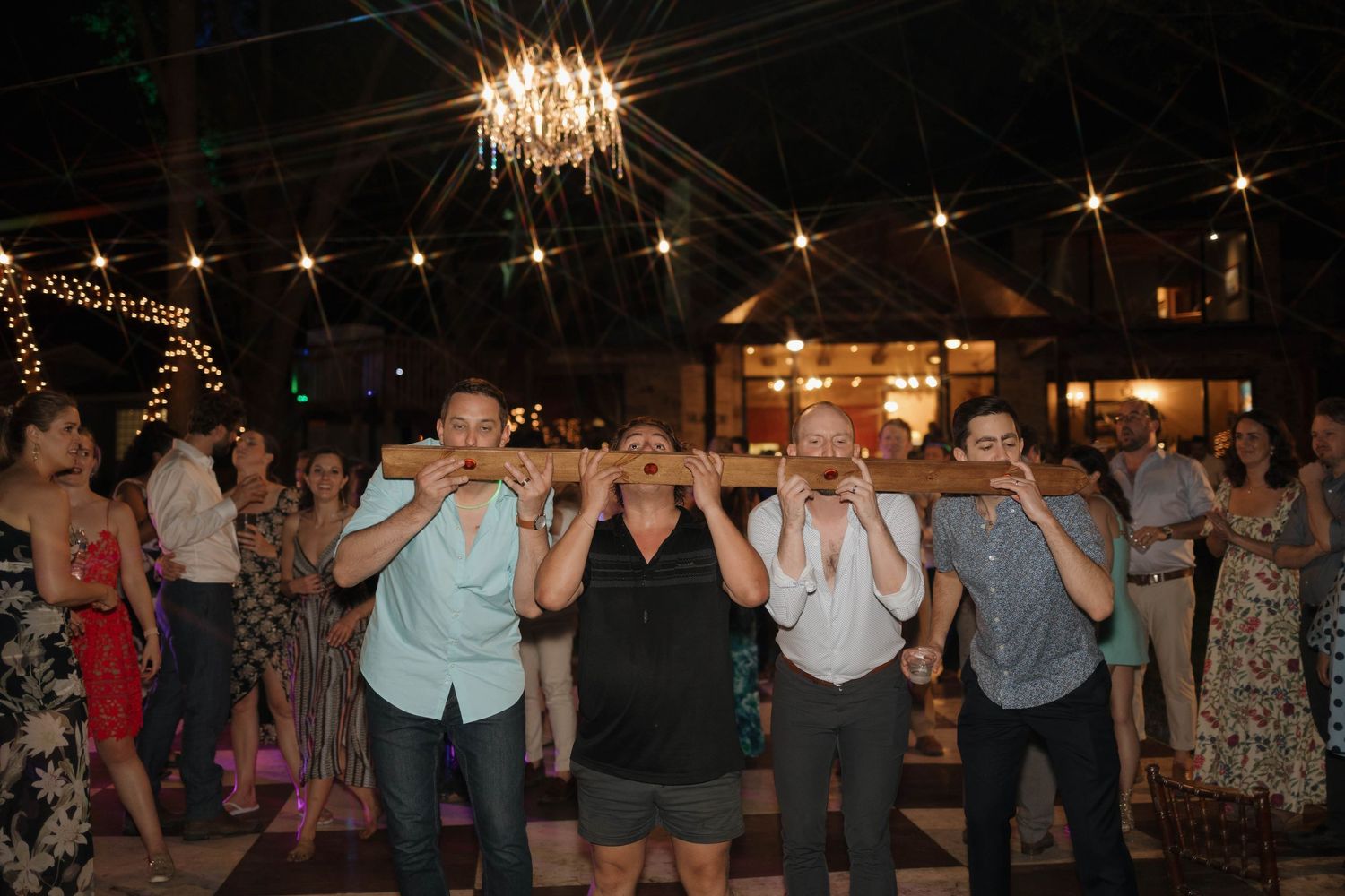 Party guests dance together under twinkling string lights and a chandelier at an outdoor evening celebration.