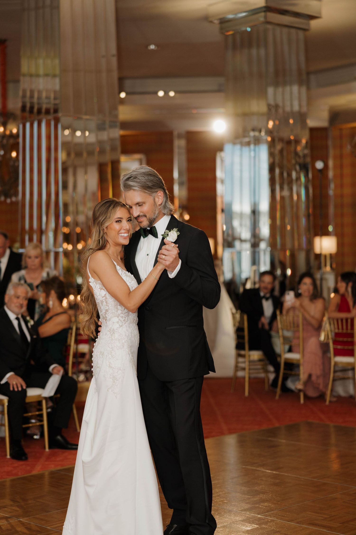 A couple shares their first dance in an elegant ballroom with mirrored columns and soft lighting during their wedding reception.