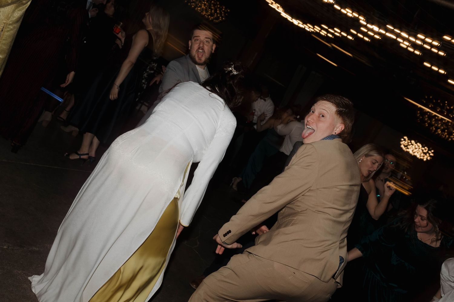 Two people dancing at an event, with one wearing a long white dress and another in a tan suit.