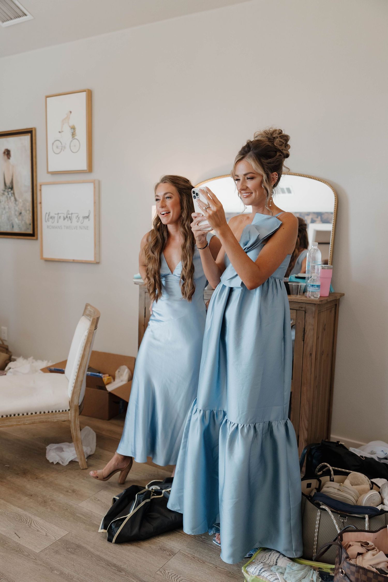 Two friends in matching light blue dresses laugh together while getting ready in a bright room with framed artwork.