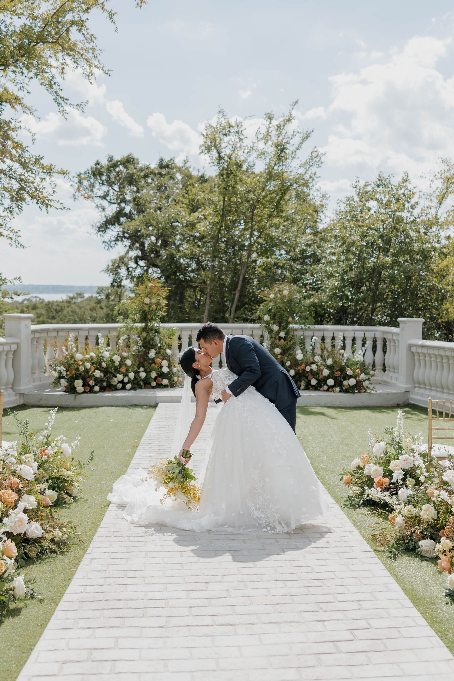 A couple shares a romantic dip kiss on their wedding day on an elegant terrace decorated with floral arrangements.