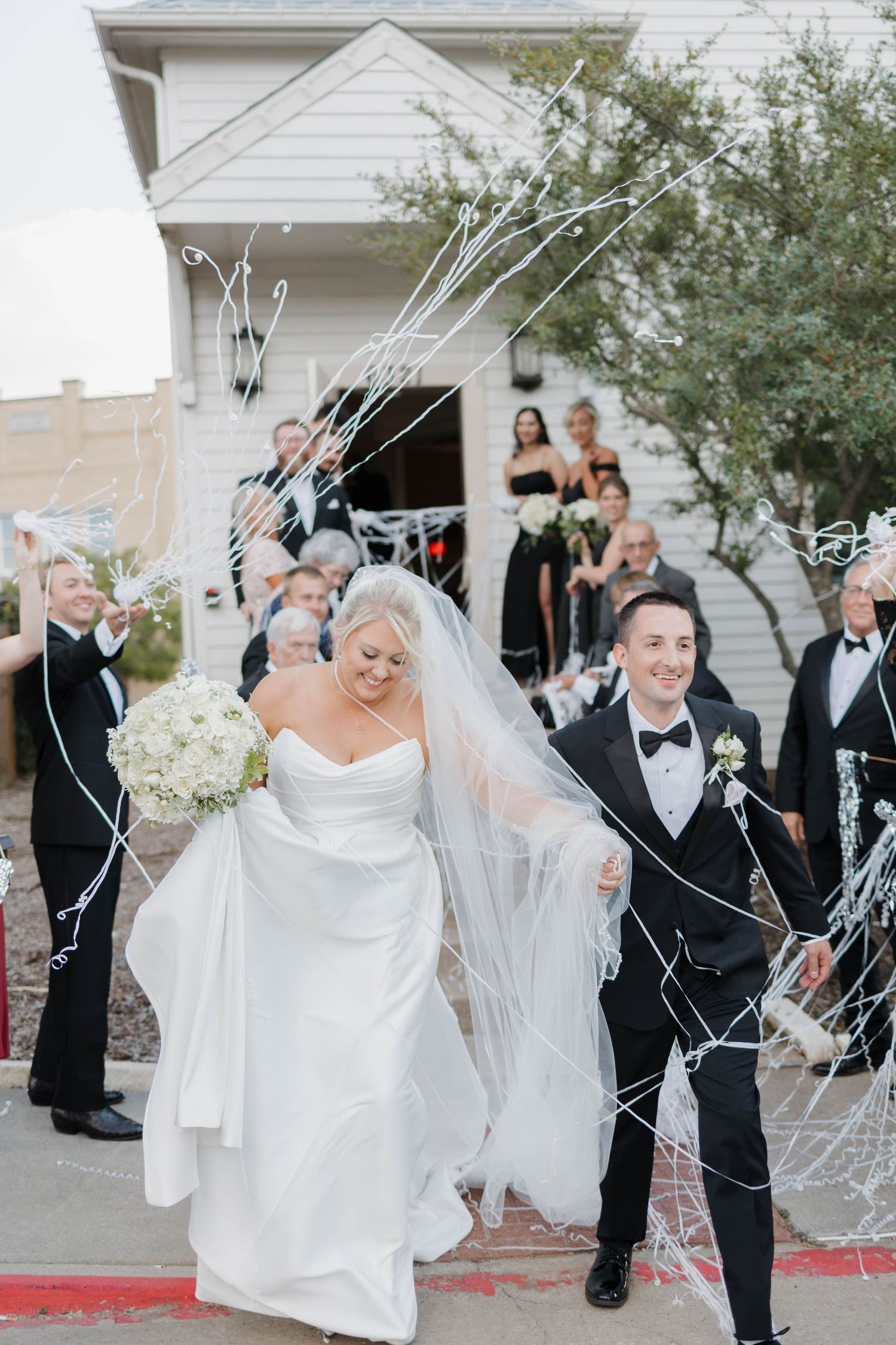 Wedding exit celebration with guests tossing white streamers as newlyweds walk through a decorated garden pathway.