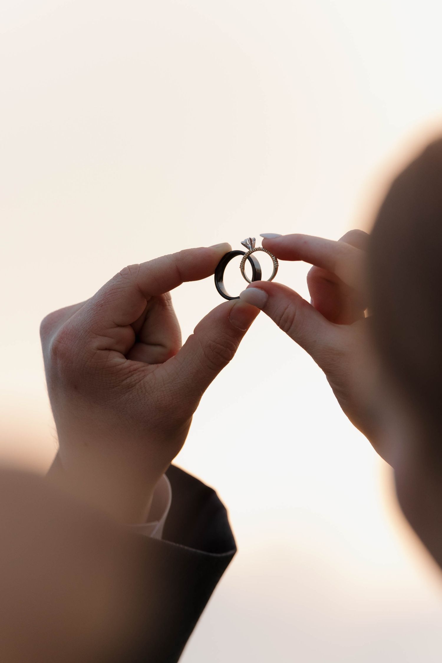 Two hands hold an engagement ring up to the sky during sunset, creating a silhouetted romantic scene.