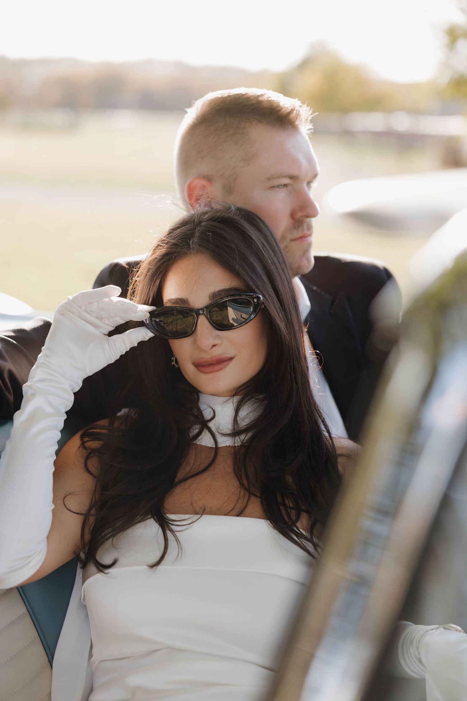 A couple in formal attire pose together outdoors, with the person in white wearing sunglasses and long gloves.