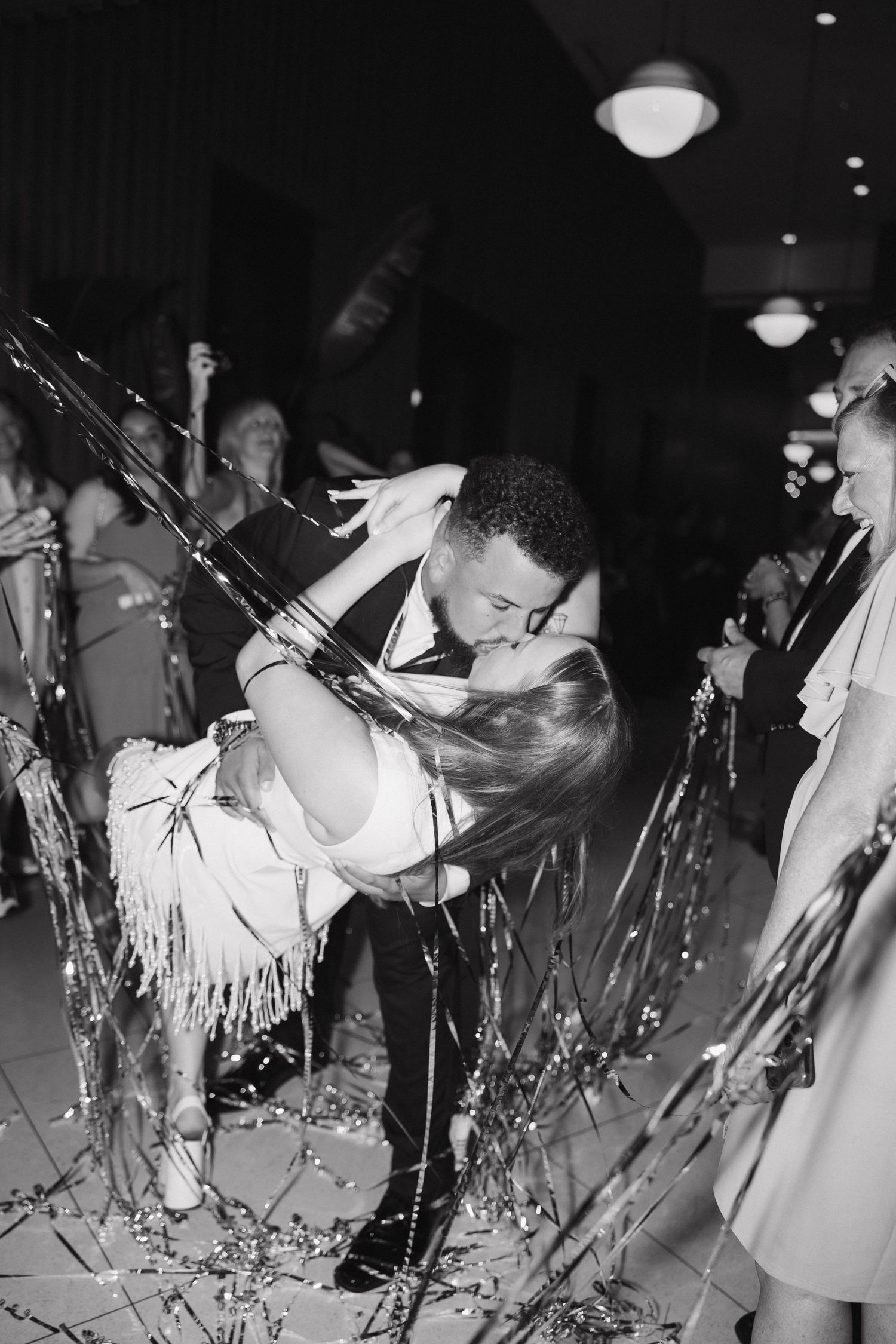 A romantic couple shares a New Year's Eve kiss at a party surrounded by streamers and glittering decorations.