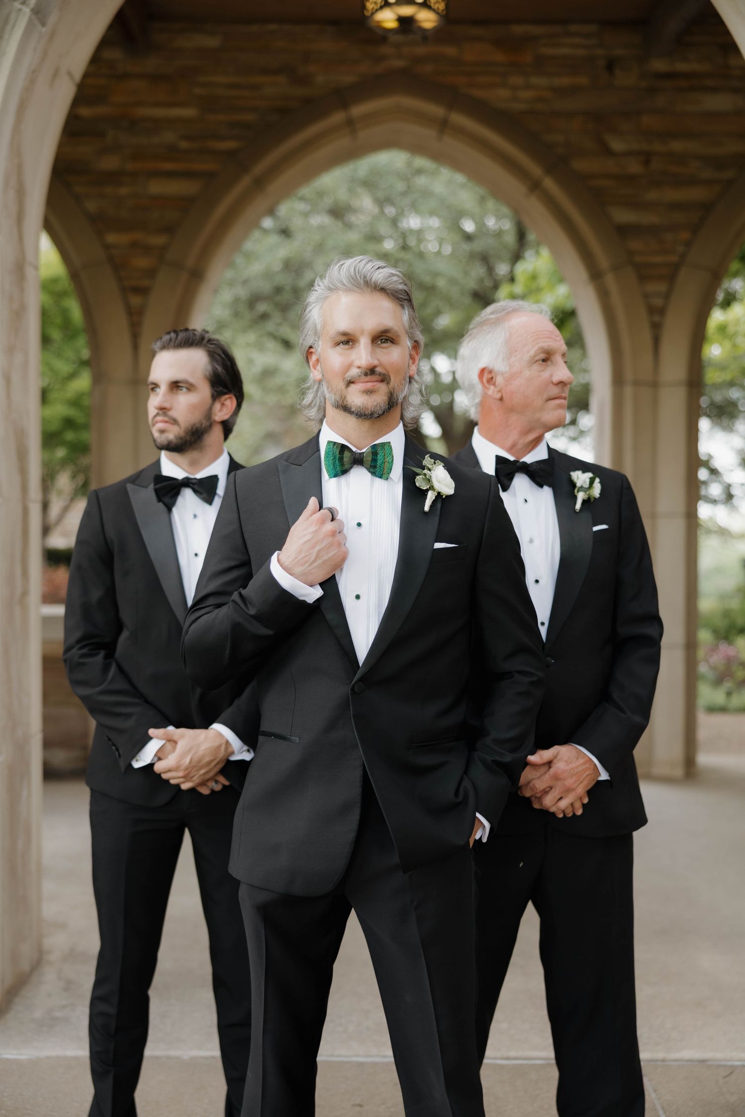 Groomsmen in classic black tuxedos and bow ties pose together under an arched colonnade.