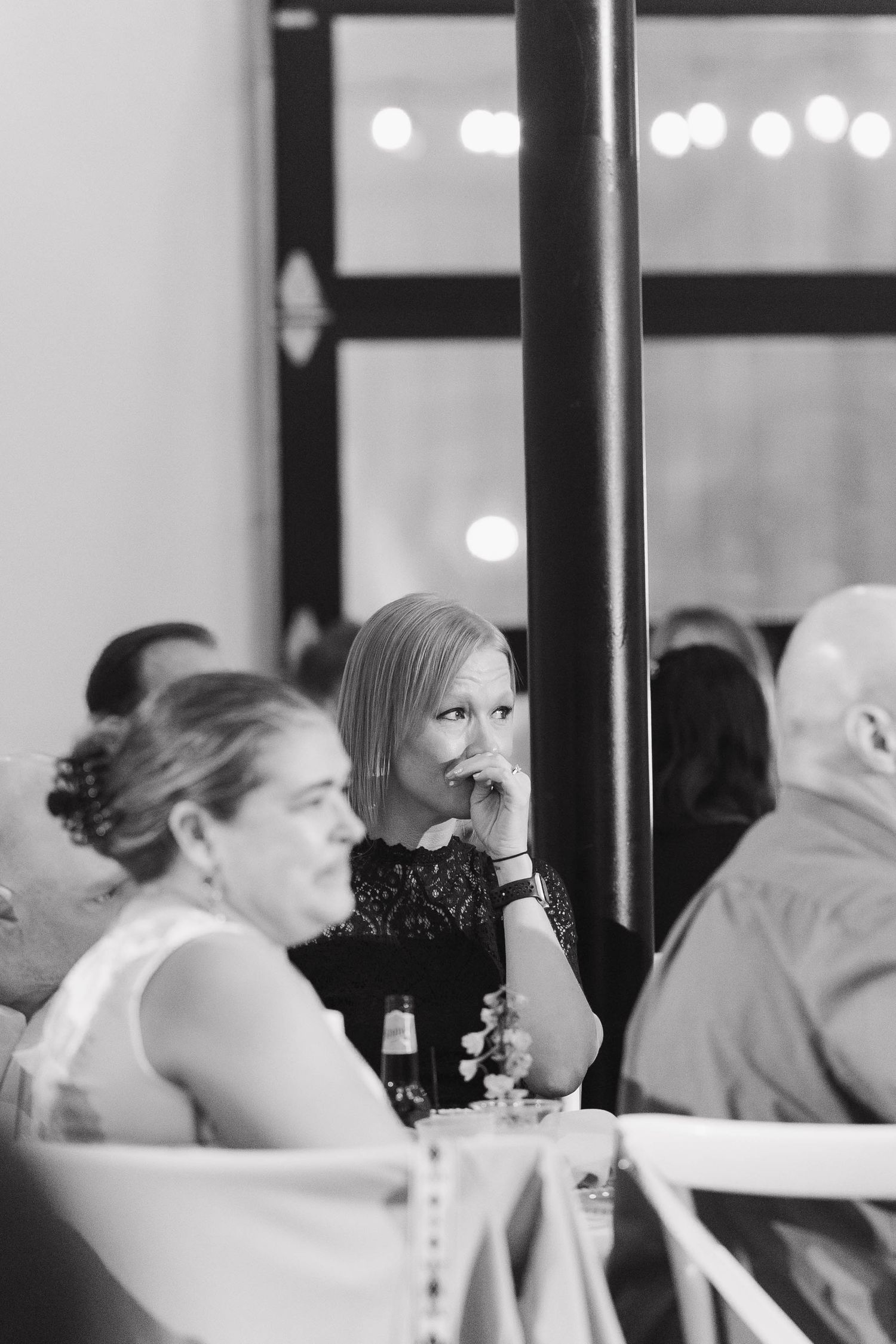 Black and white candid photo showing wedding guests seated at tables with glowing bokeh lights in the background.