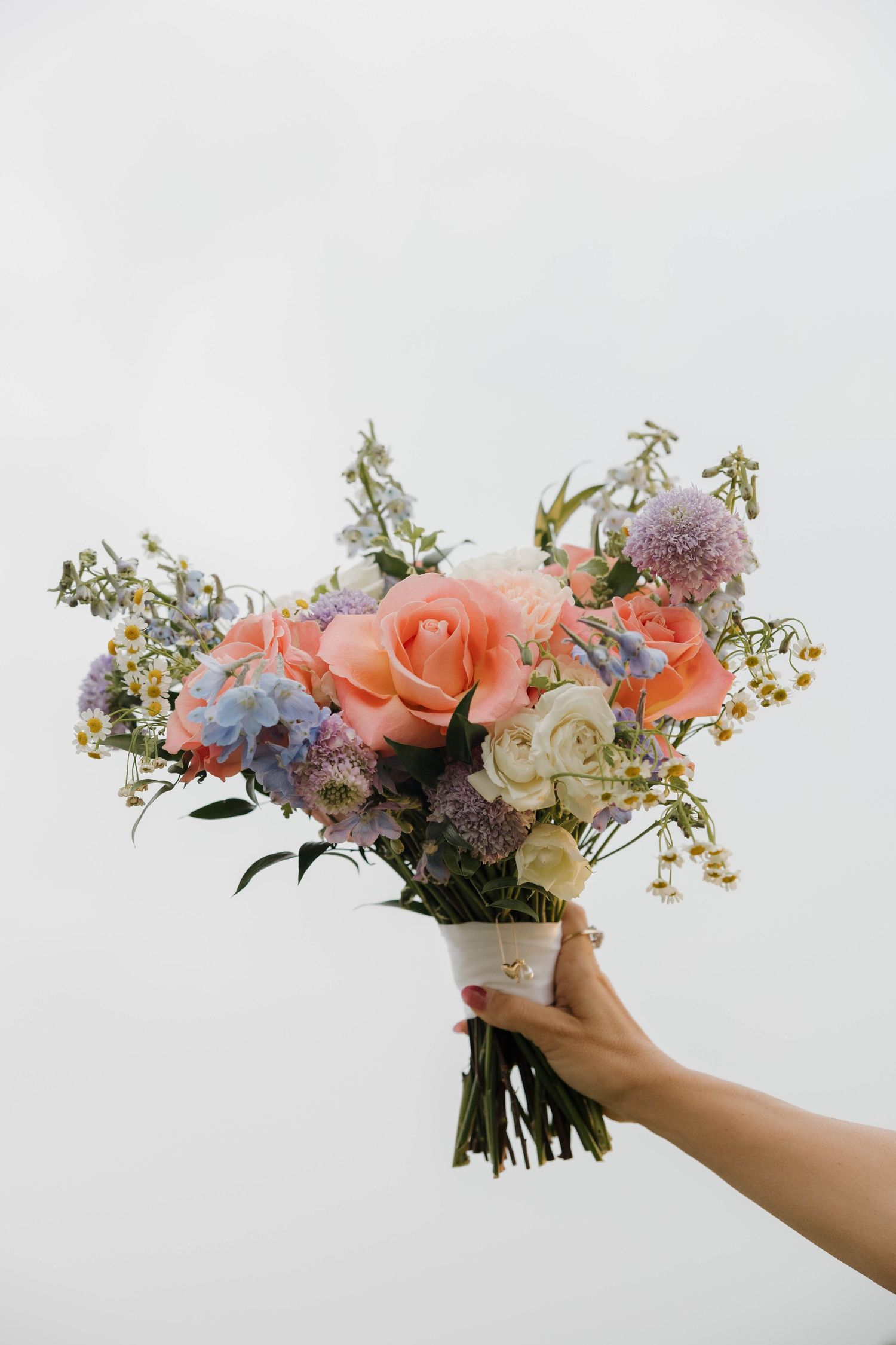 Hand holding a romantic bouquet with peach roses, purple flowers and delicate greenery against a white background.