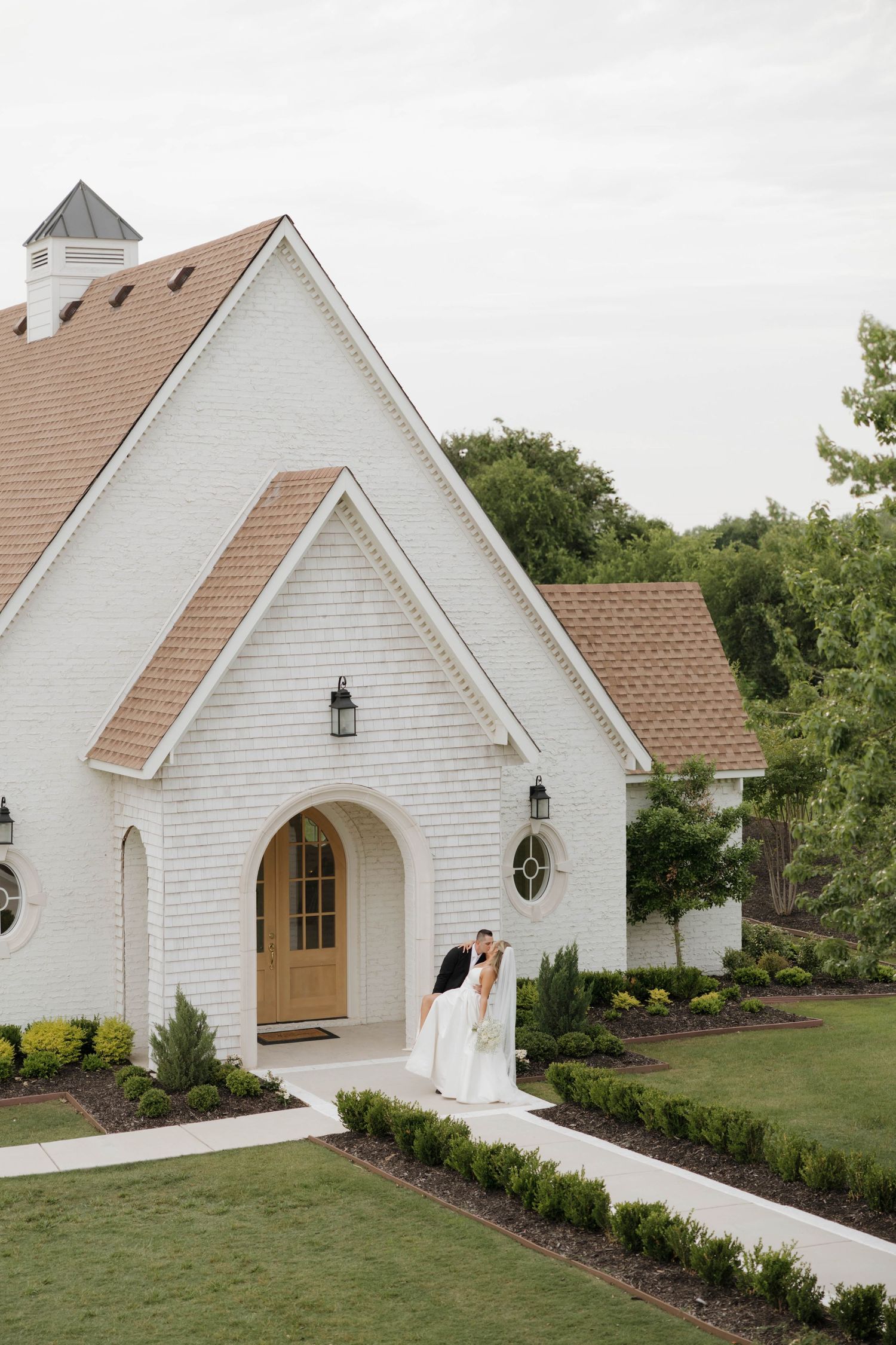 A charming white chapel with tan accents and a tower features an arched wooden door entrance surrounded by manicured landscaping.