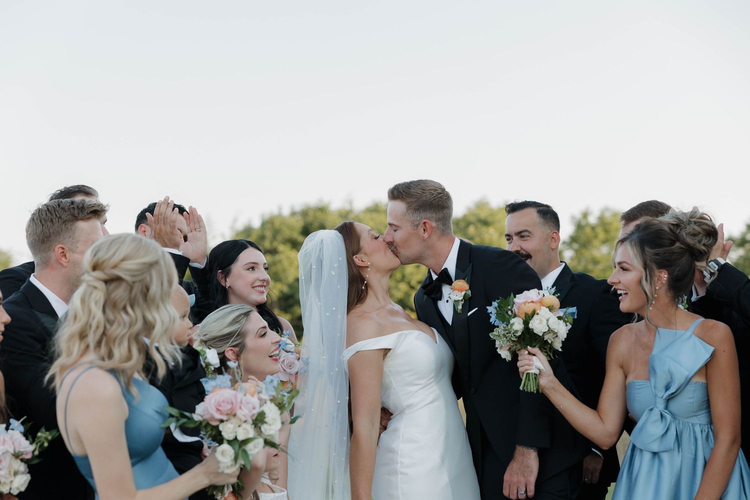 A loving couple shares an affectionate moment with their wedding party in light blue dresses and suits in the background.