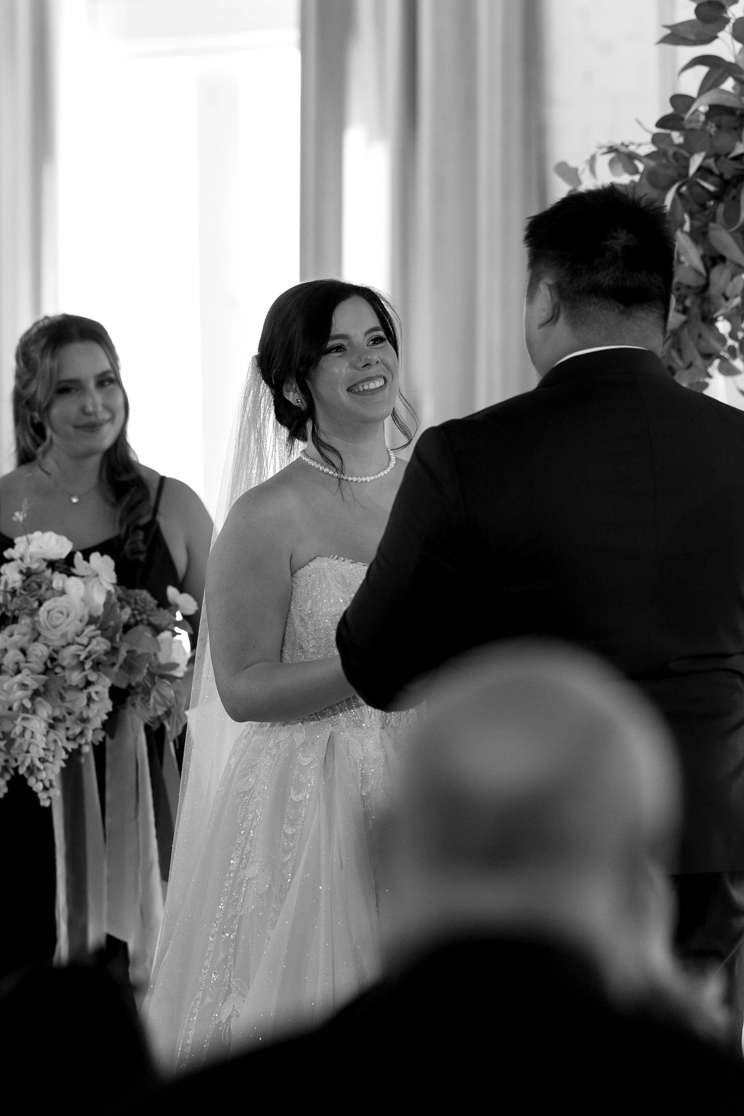 A joyful moment during a wedding ceremony as bride smiles brightly while exchanging vows with white floral decor nearby.