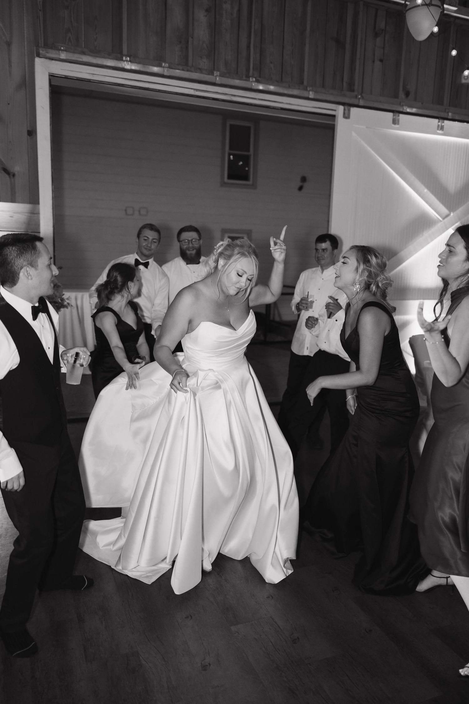 A bride in a strapless white ballgown dances joyfully on the dance floor surrounded by wedding guests at a reception.