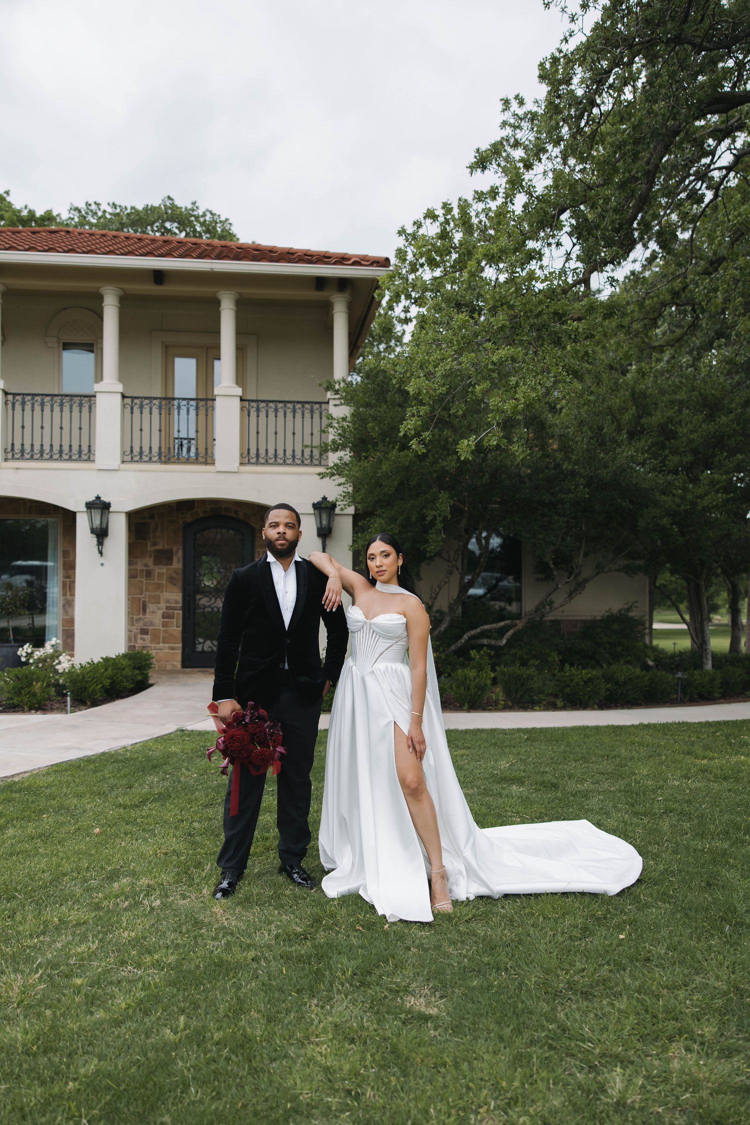 Newlyweds pose in front of a Mediterranean-style villa with a white wedding dress and deep red bouquet.