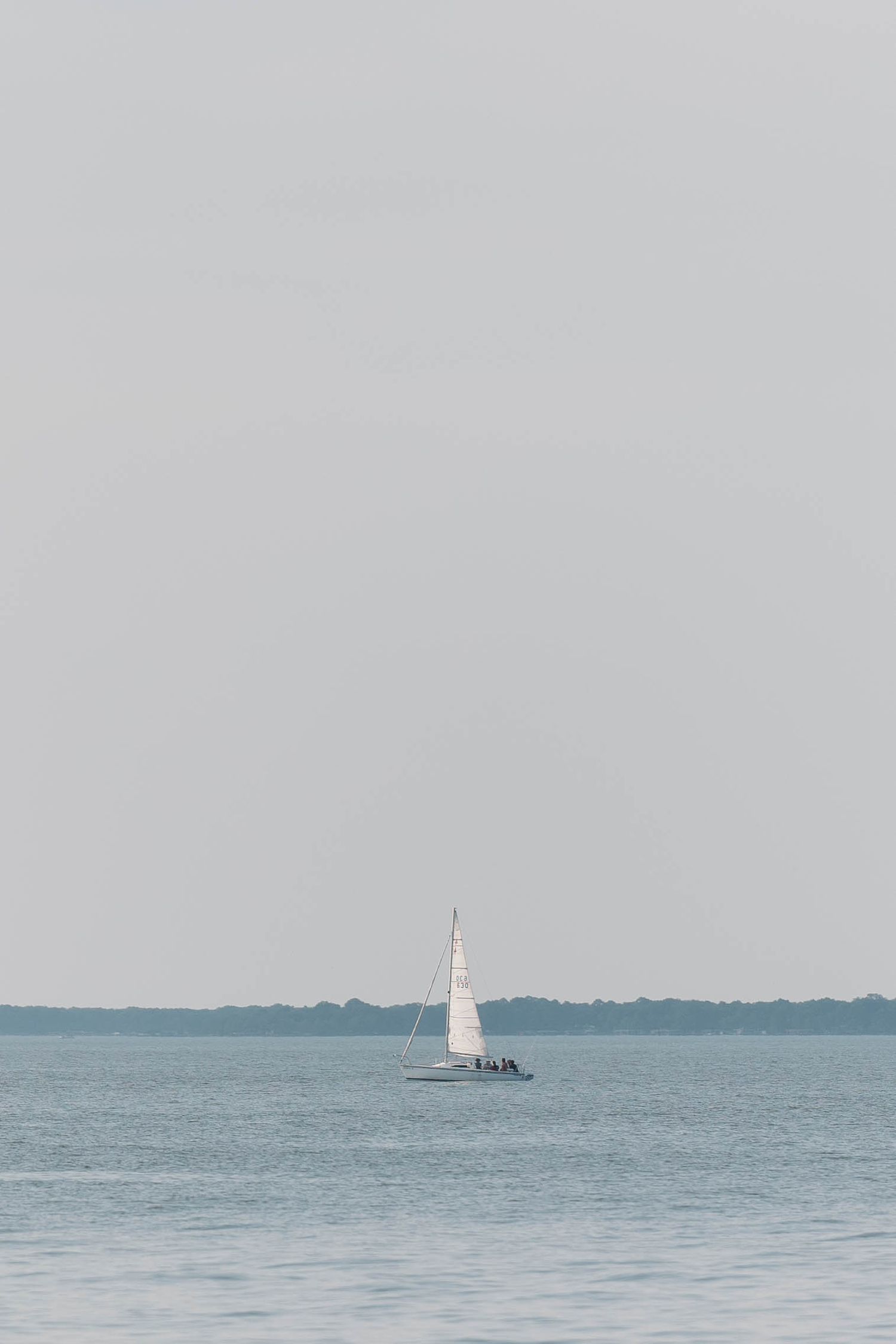 A small white sailboat glides across calm blue waters under an overcast sky in a minimalist seascape.
