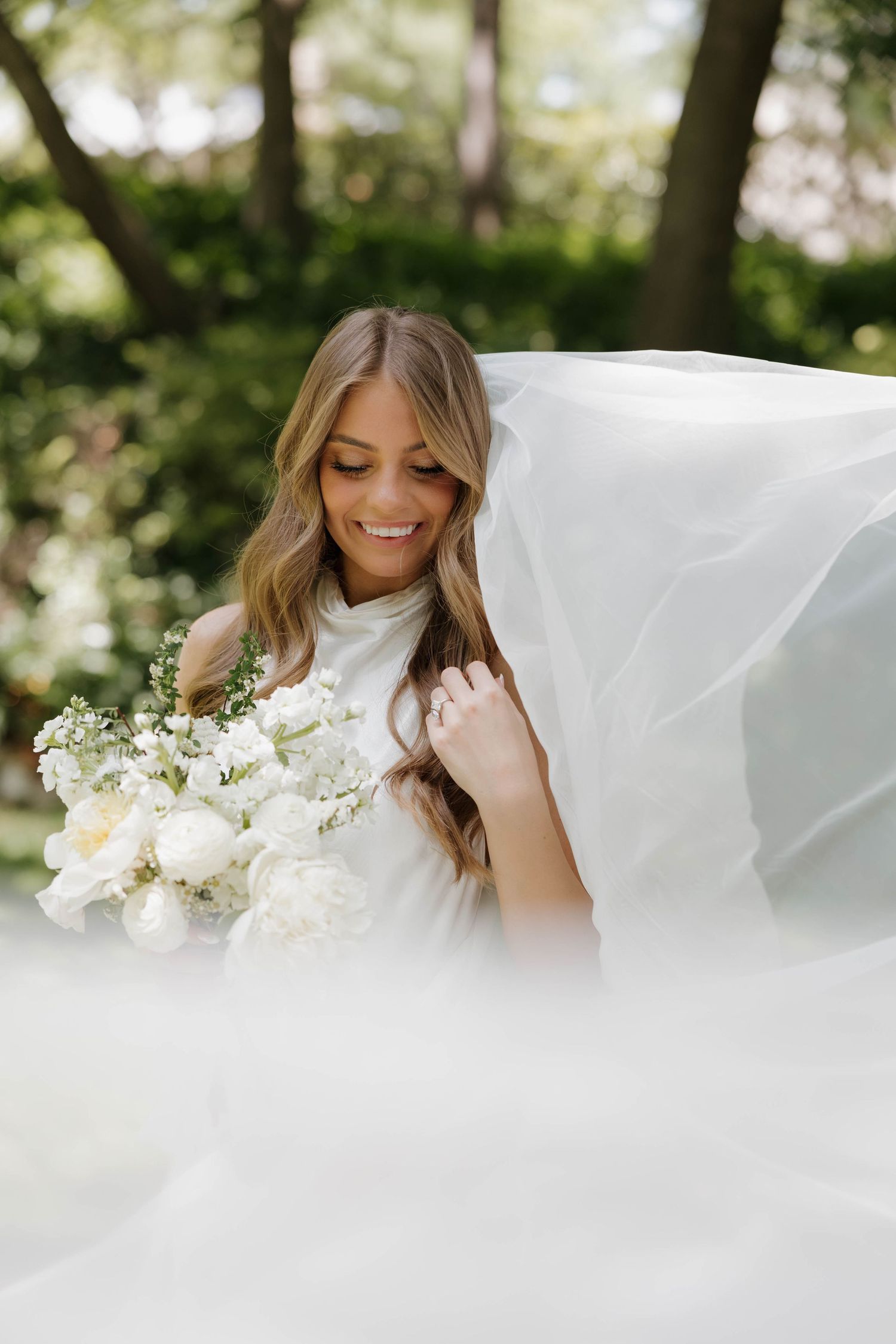 Bride wearing flowy white veil holds elegant white bouquet while smiling in outdoor garden setting with bokeh effect.