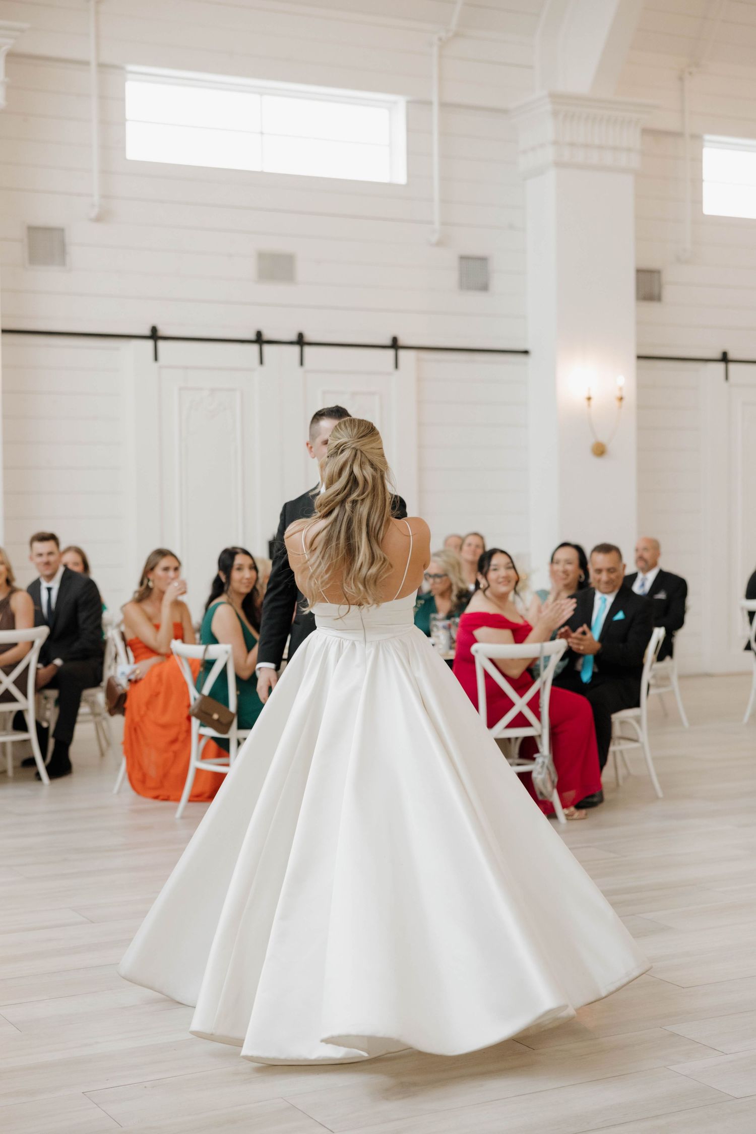 A wedding celebration in a bright white modern venue with guests seated in cross-back chairs watching a special moment.