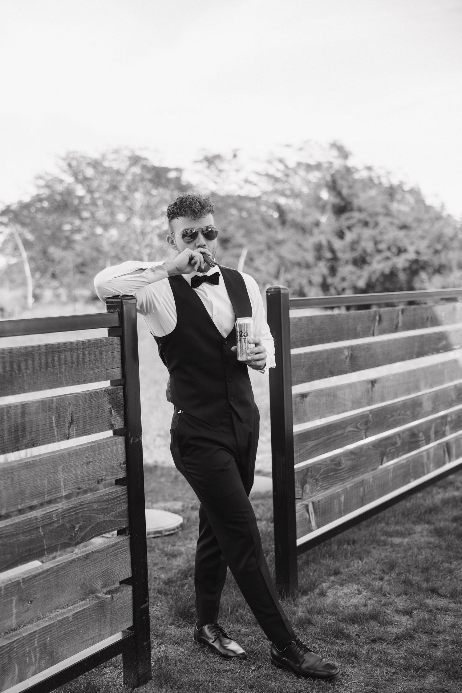 A person in formal attire with sunglasses and bowtie leans against a wooden fence while holding a drink.