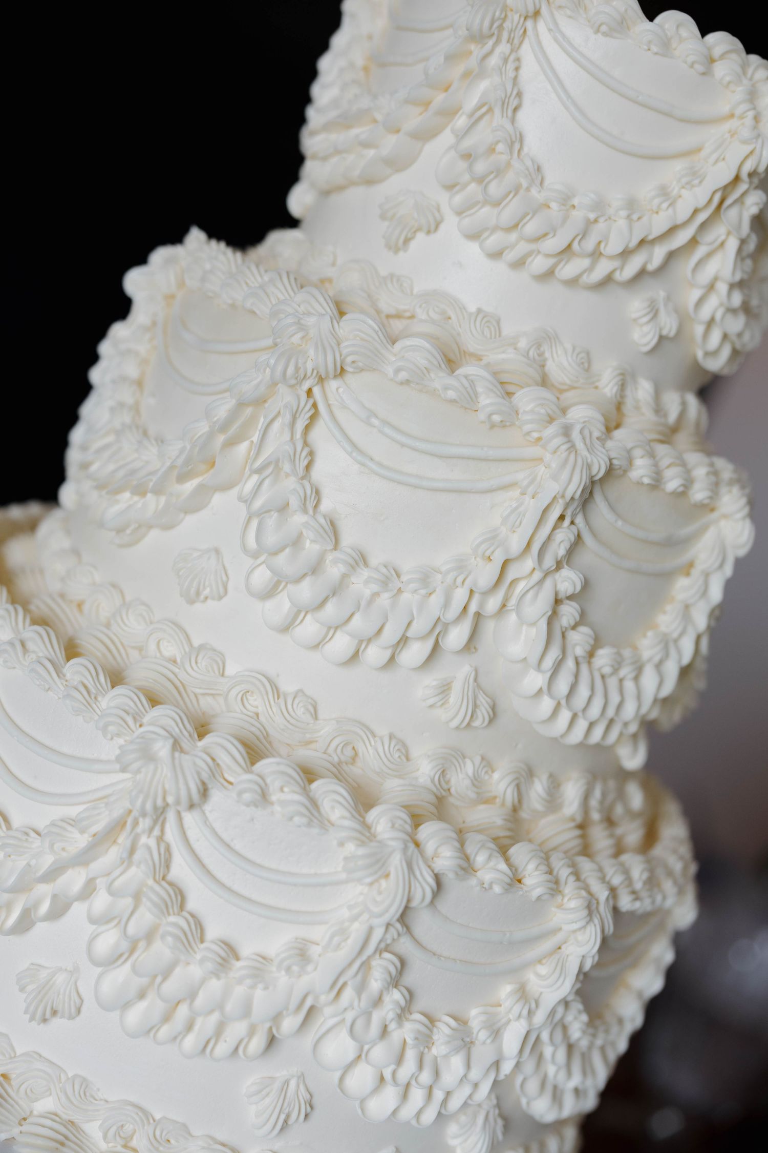 Close-up of an elegant white wedding cake with ornate piping detail featuring scalloped designs and delicate lace patterns.