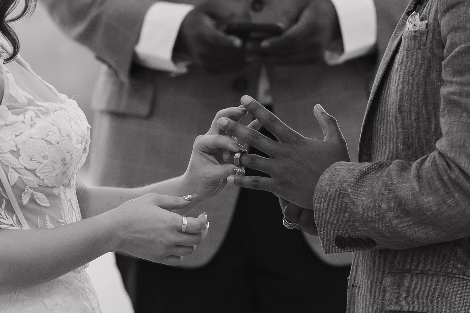 Black and white image of two hands exchanging rings during a wedding ceremony, with formal attire visible.