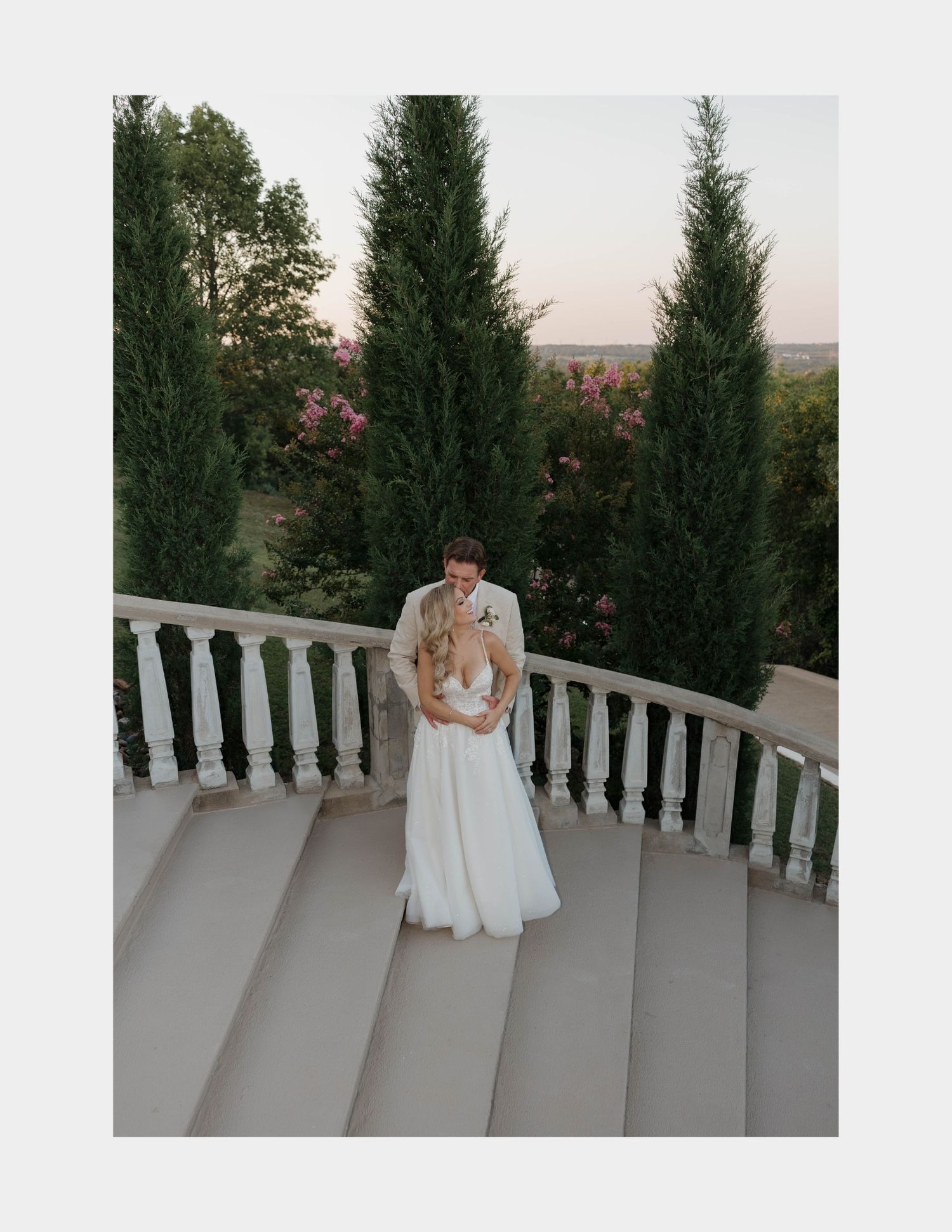 A romantic moment captured between a couple in wedding attire on an elegant white balcony surrounded by evergreen trees.