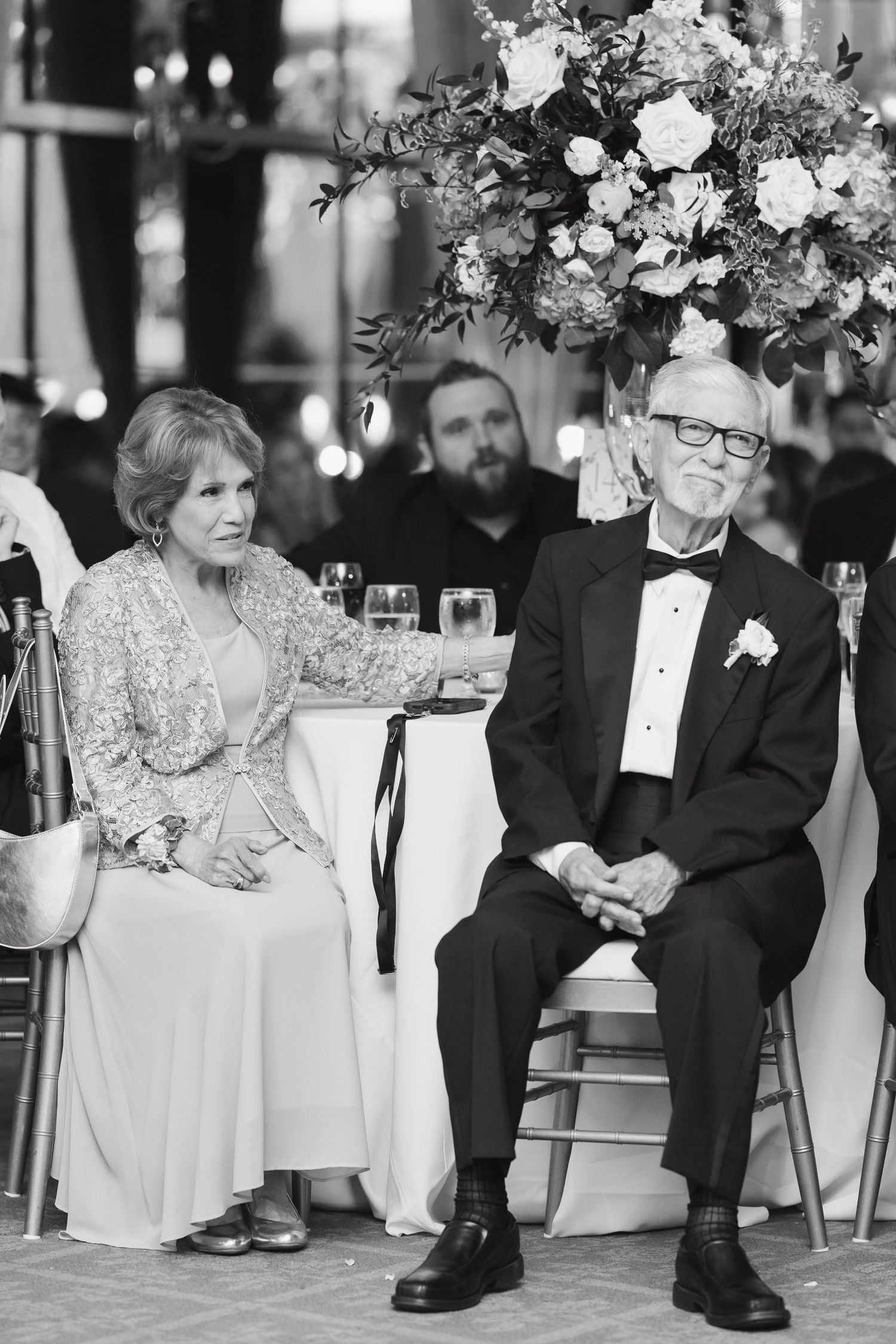 Black and white photo of formal wedding guests seated at an ornate reception dinner with floral centerpiece.