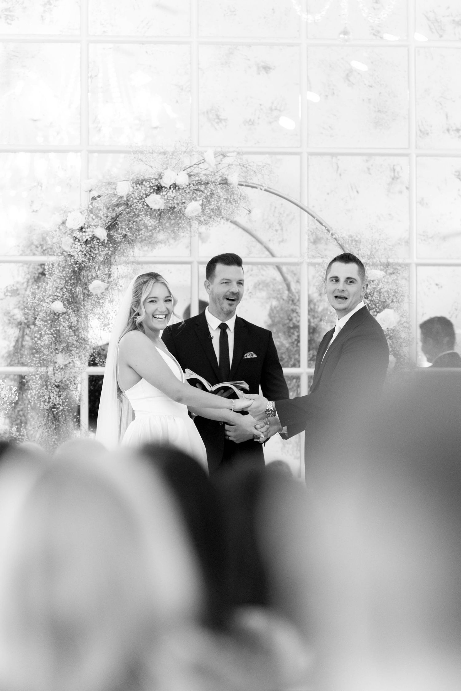 A black and white wedding ceremony photo under a floral arch with guests in attendance during an outdoor celebration.