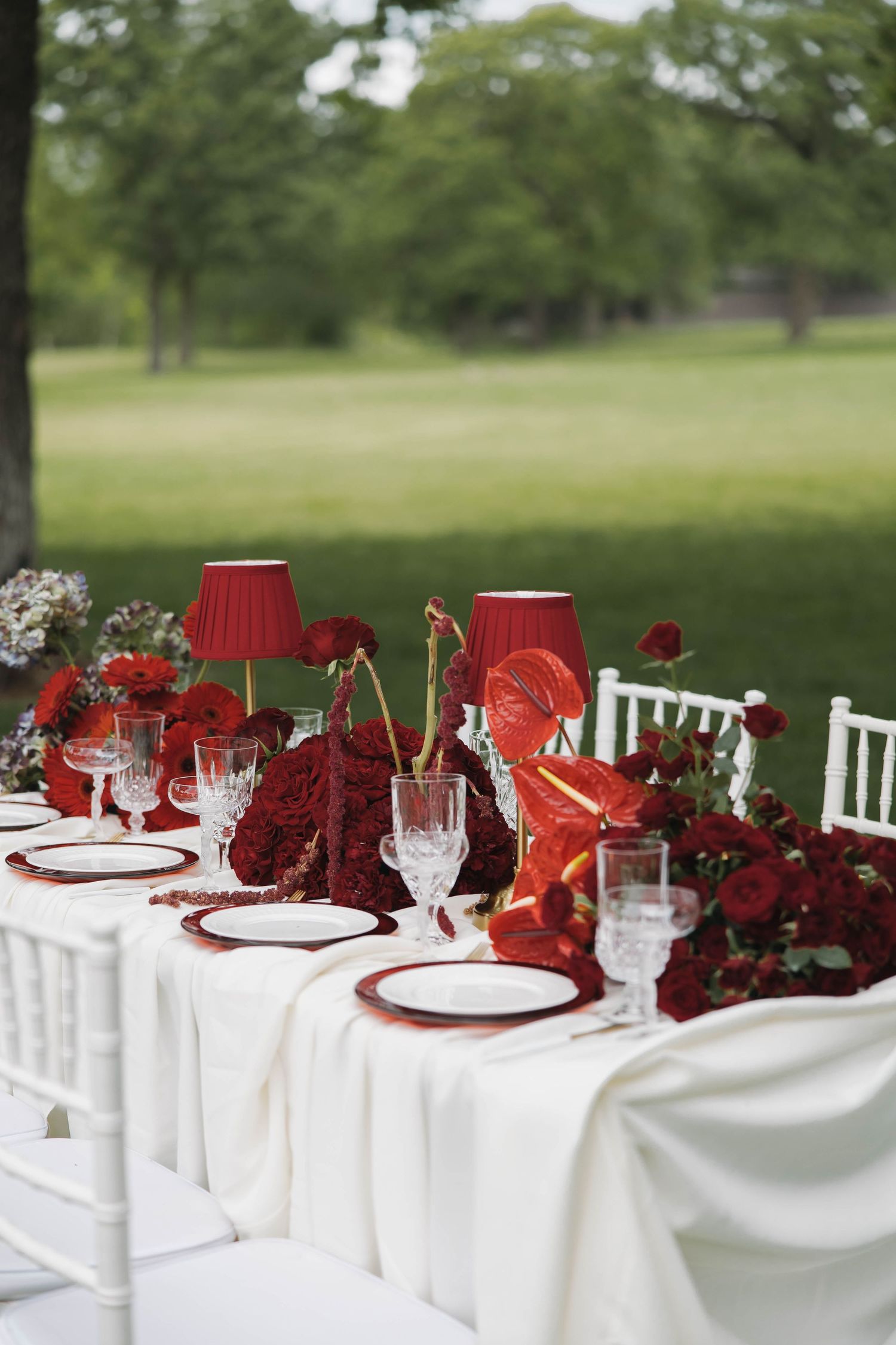 Elegant outdoor table setting with red and white decor featuring red lamps, roses, and white tablecloth and chairs.