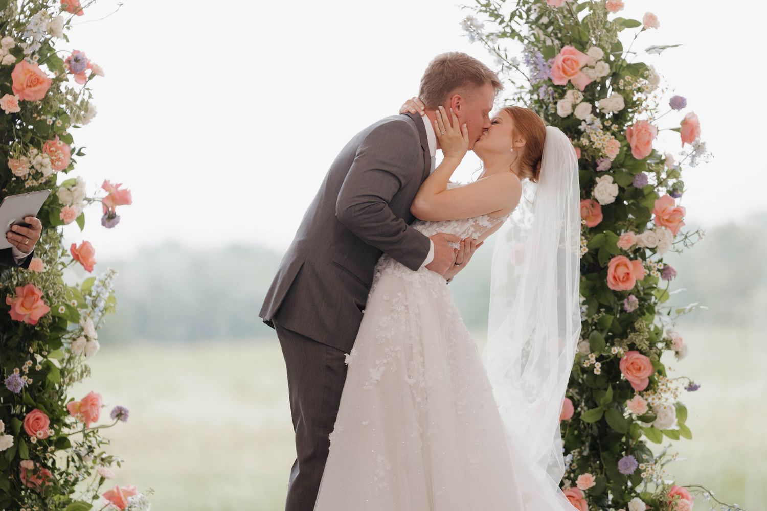 A couple shares a tender moment under a floral arch adorned with pink and white roses during an outdoor ceremony.
