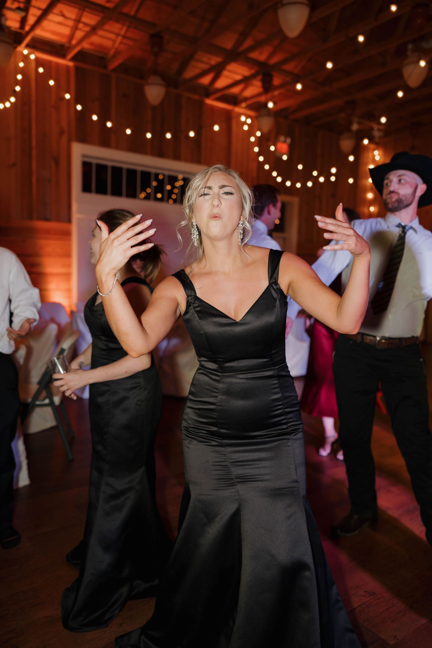 Wedding guests dancing energetically in a rustic barn venue decorated with warm string lights and dark wood beams.