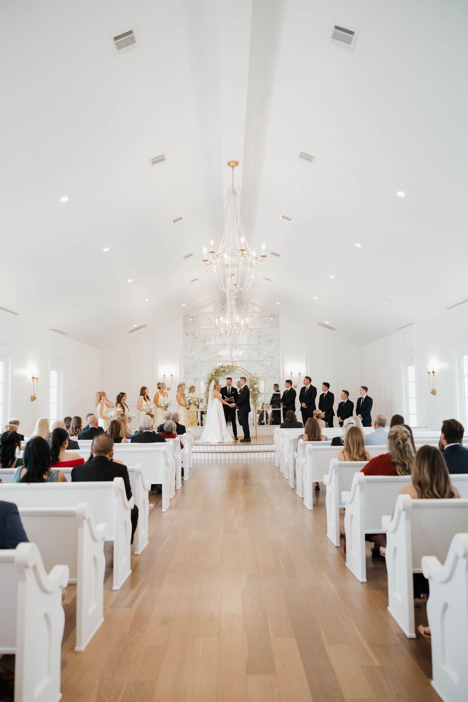 Modern white chapel interior with elegant chandelier and pews filled with wedding guests during ceremony.