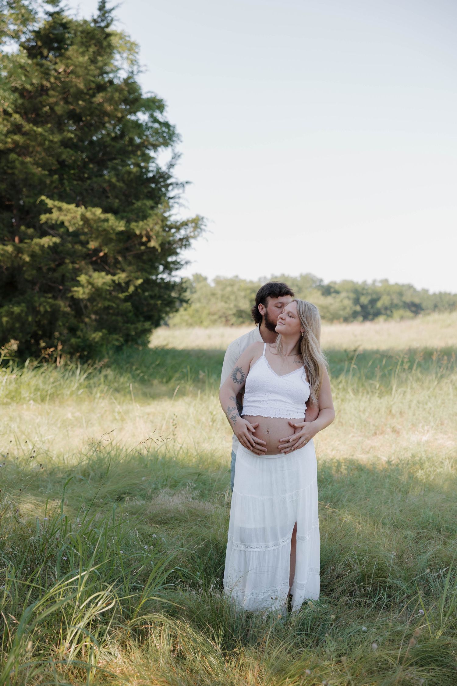 Ethereal maternity photo in white dress amid sunlit field with evergreen trees.