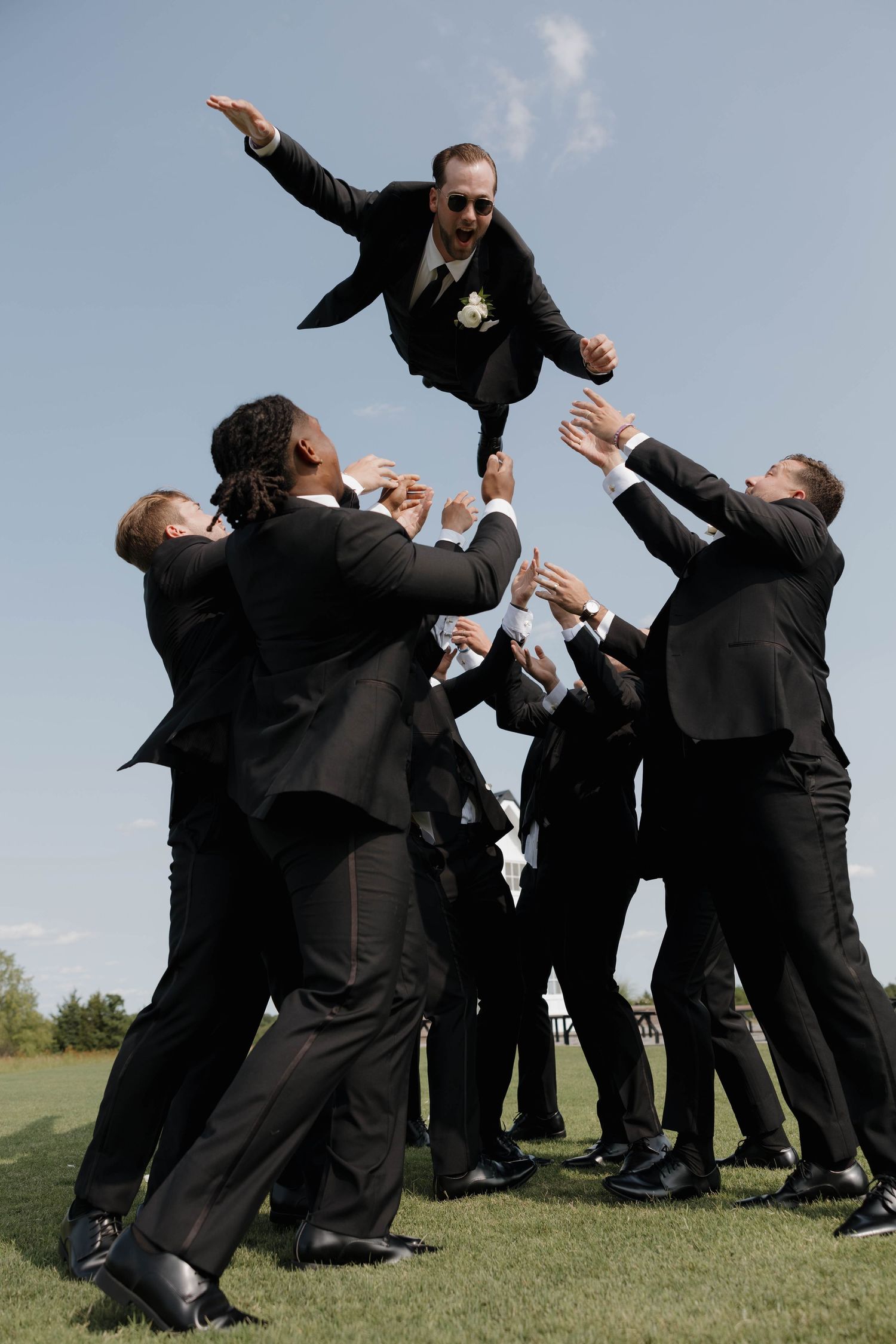 Groomsmen in black suits toss groom into air during outdoor wedding celebration photo on grassy field.